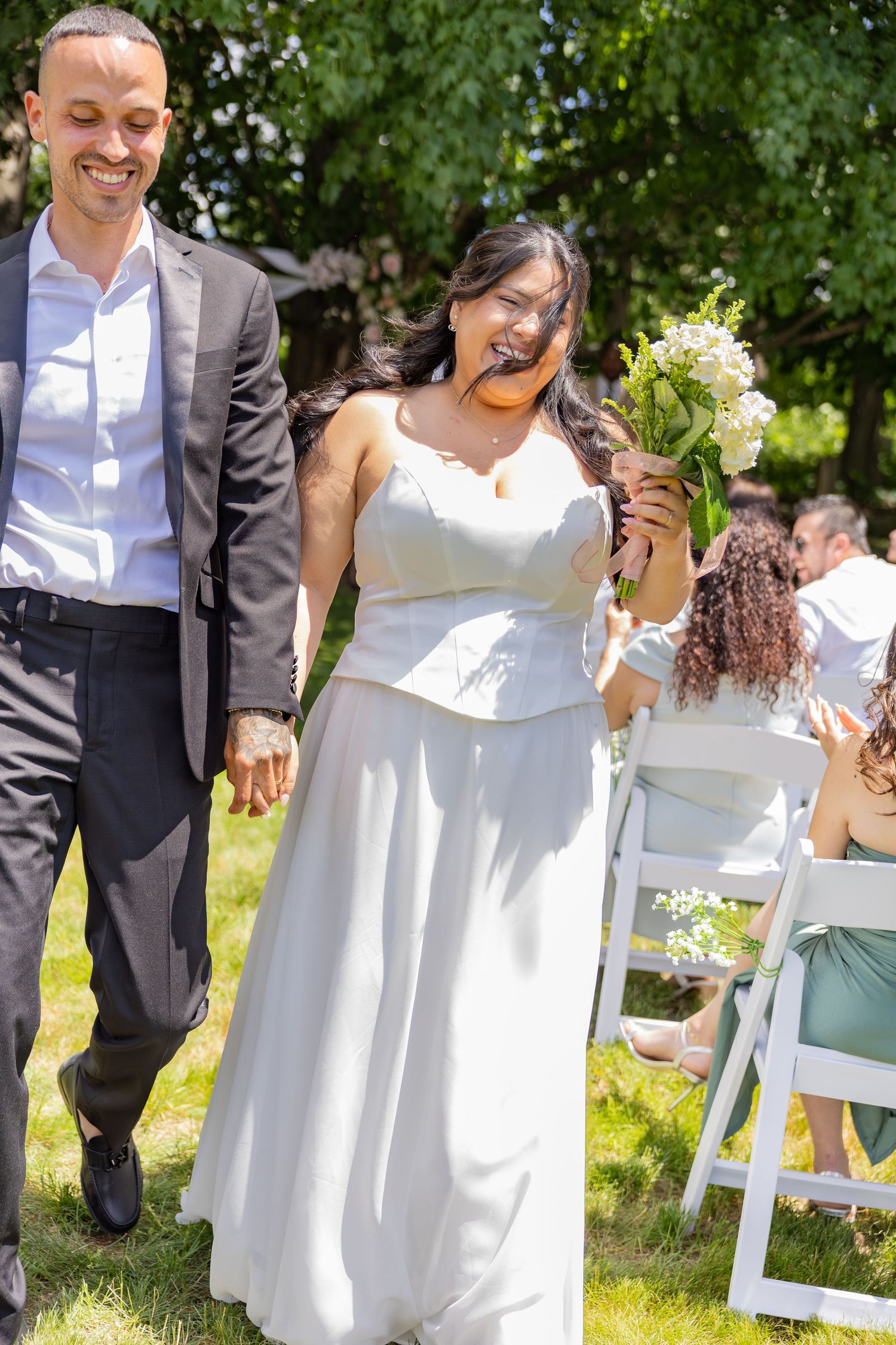 Smiling bride and groom walking hand in hand down outdoor wedding aisle in sunlight.