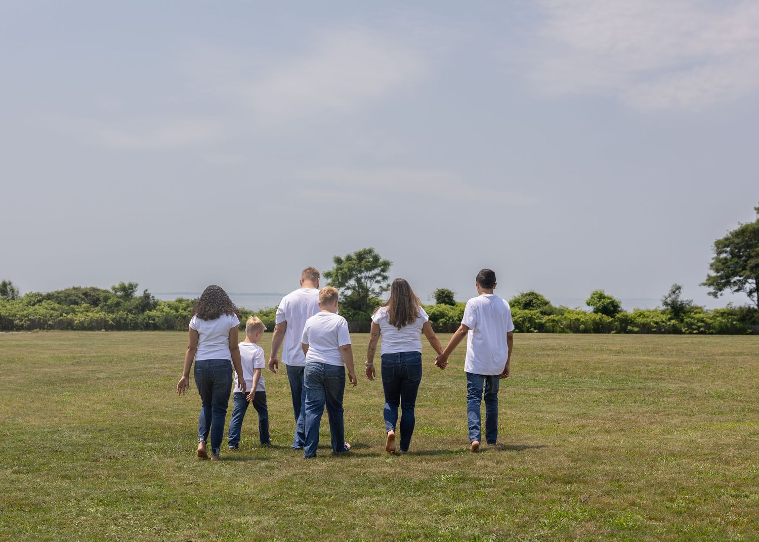 Family of six in white shirts holds hands walking across green field.