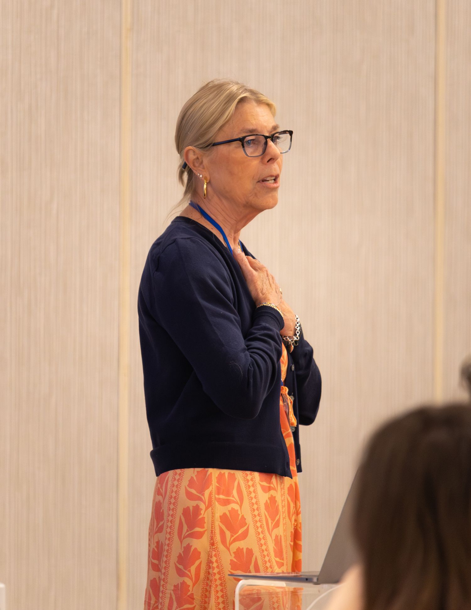 Blonde woman in navy cardigan and orange skirt speaking thoughtfully at a conference session.