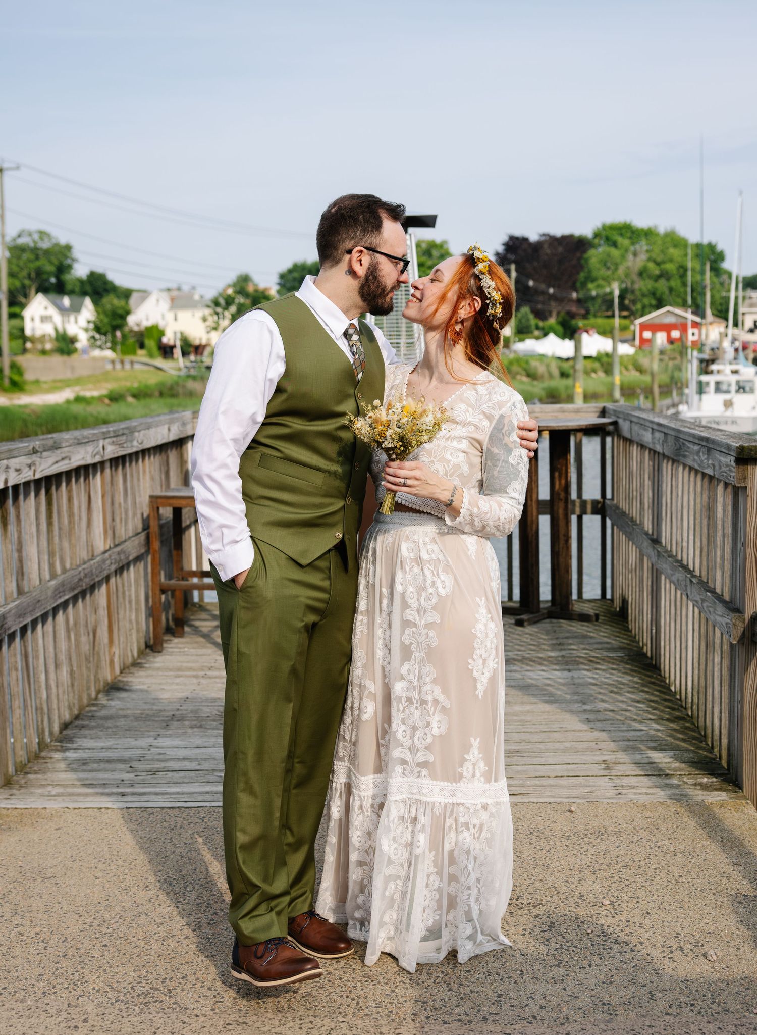 Couple in wedding attire sharing romantic moment on wooden dock with marina in background.