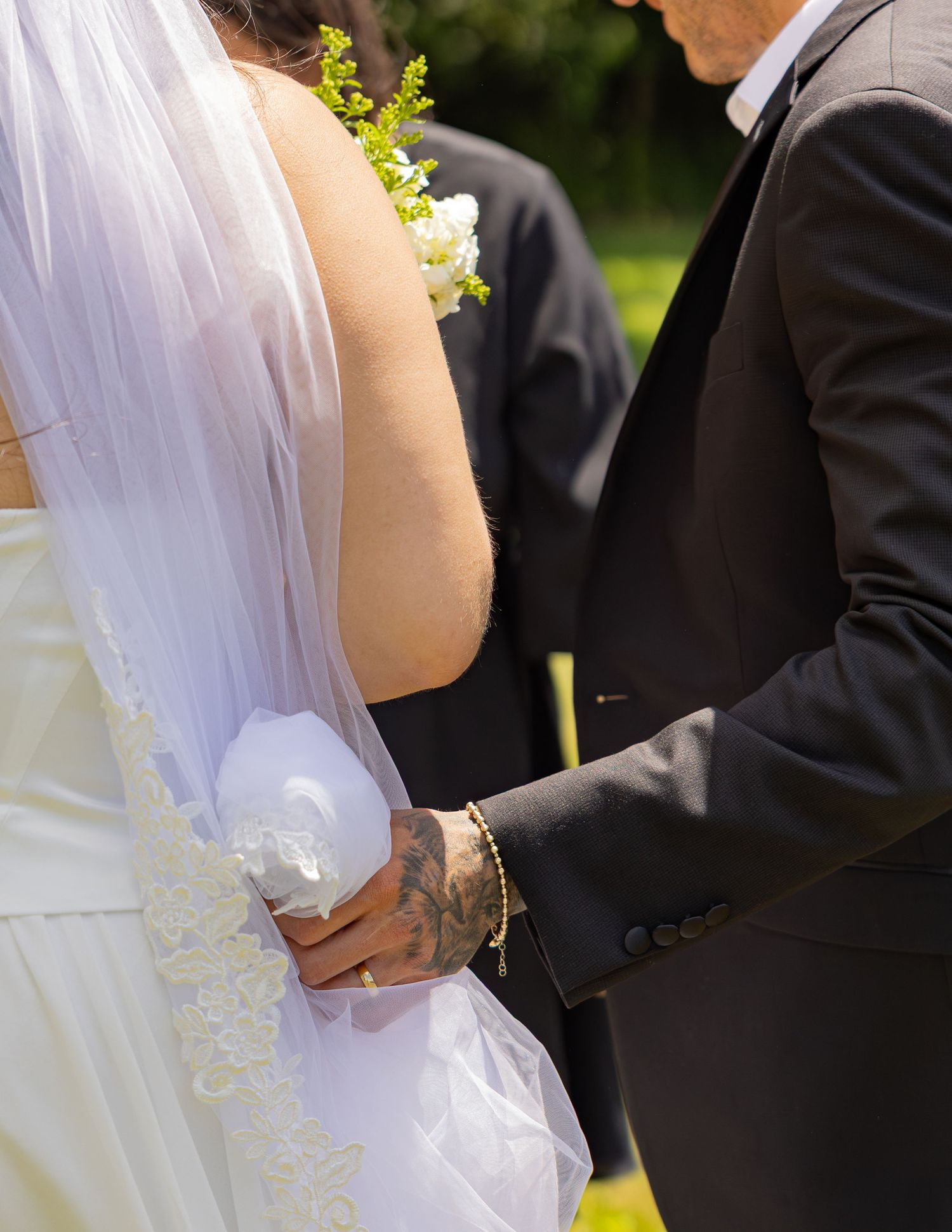 Close-up of groom holding bride's waist with tattooed hand during outdoor wedding ceremony.