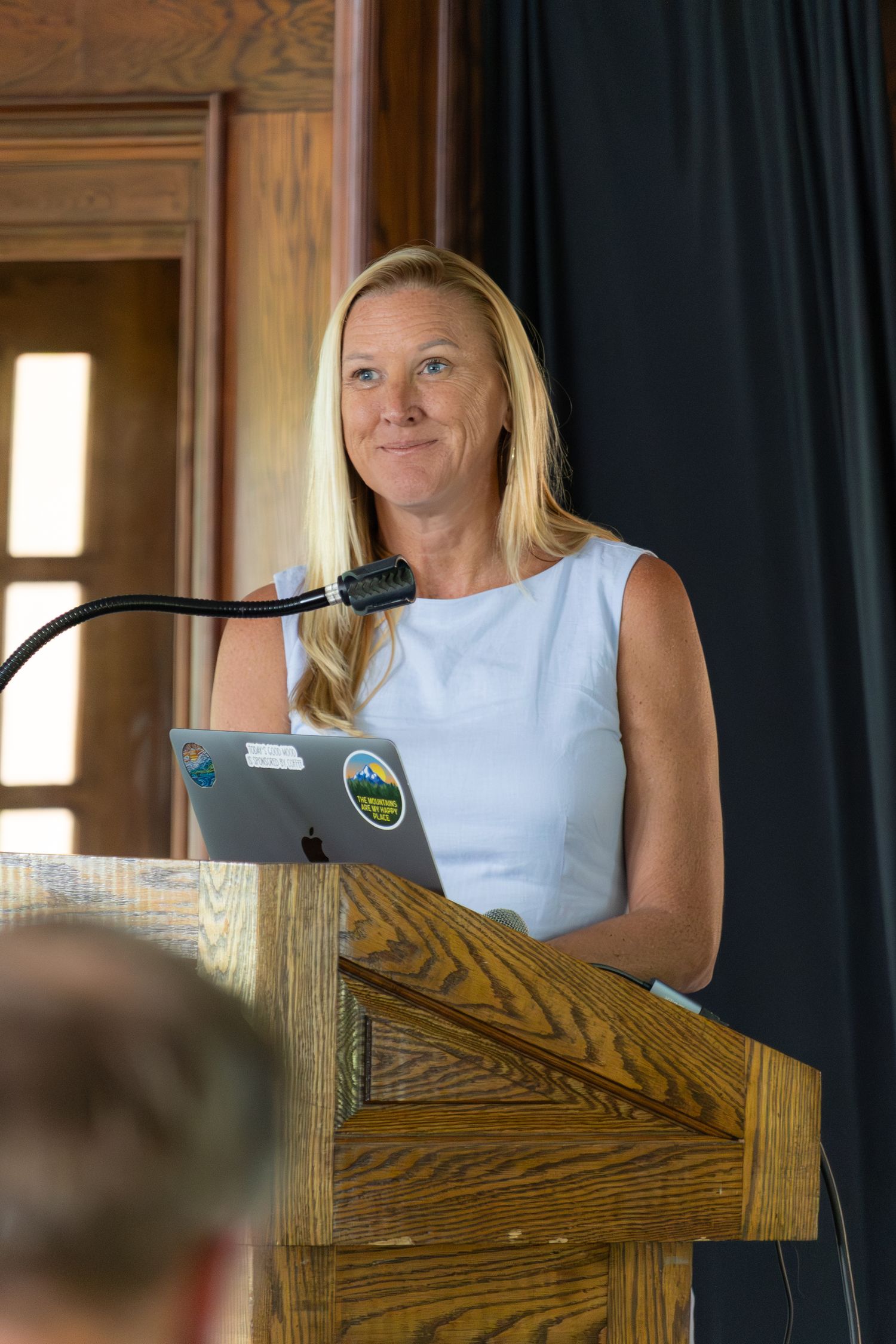 Blonde woman speaker presents at wooden podium with laptop during indoor conference event.