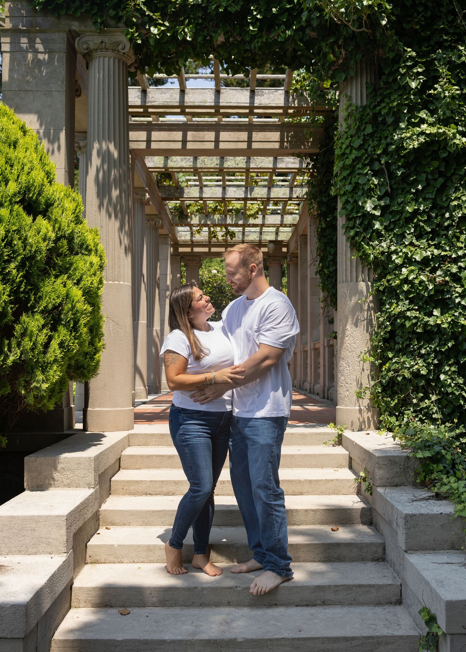 Couple embraces barefoot on stone steps under ivy-covered pergola in garden.
