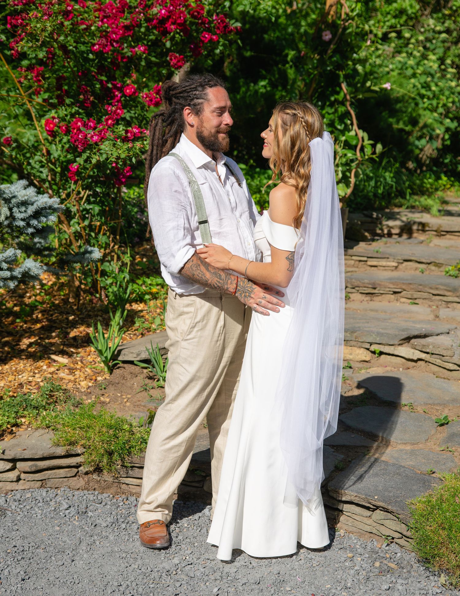 Bride in white off-shoulder gown and groom with dreadlocks embrace on stone path amid red roses.