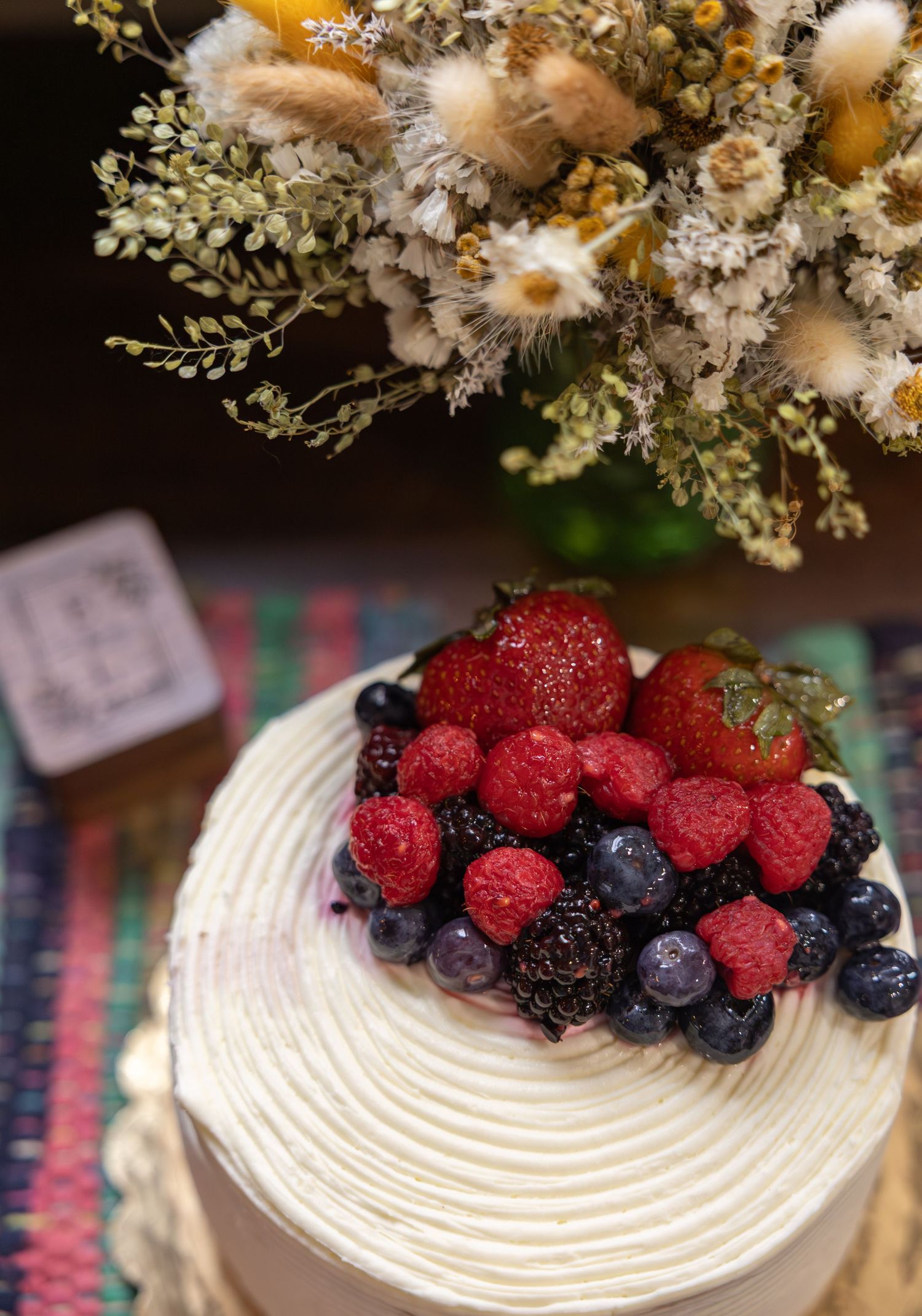 Wedding cake topped with fresh strawberries, raspberries, and blueberries beside a dried floral bouquet.