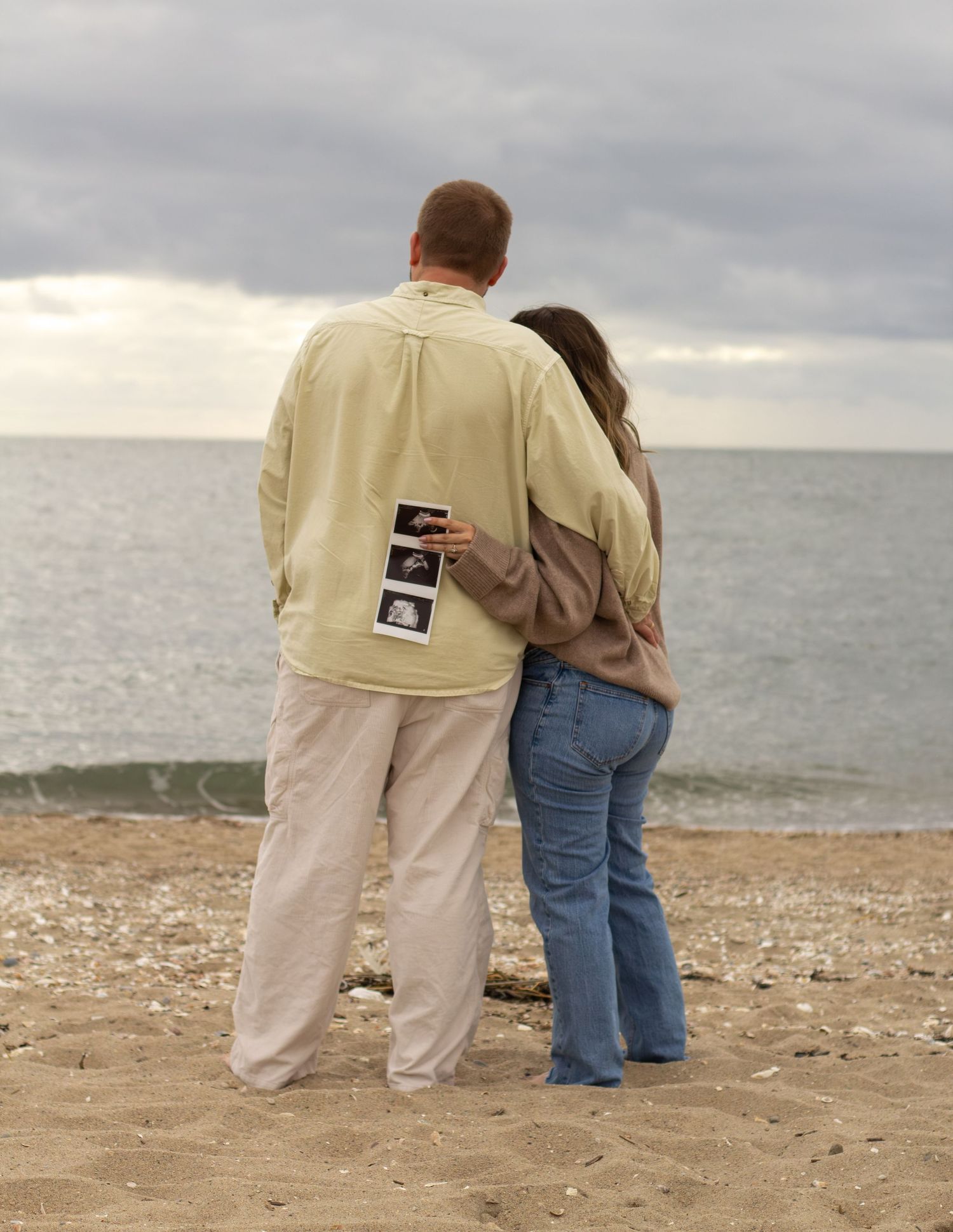 Couple standing on beach from behind, man holding ultrasound photos while embracing pregnant partner.
