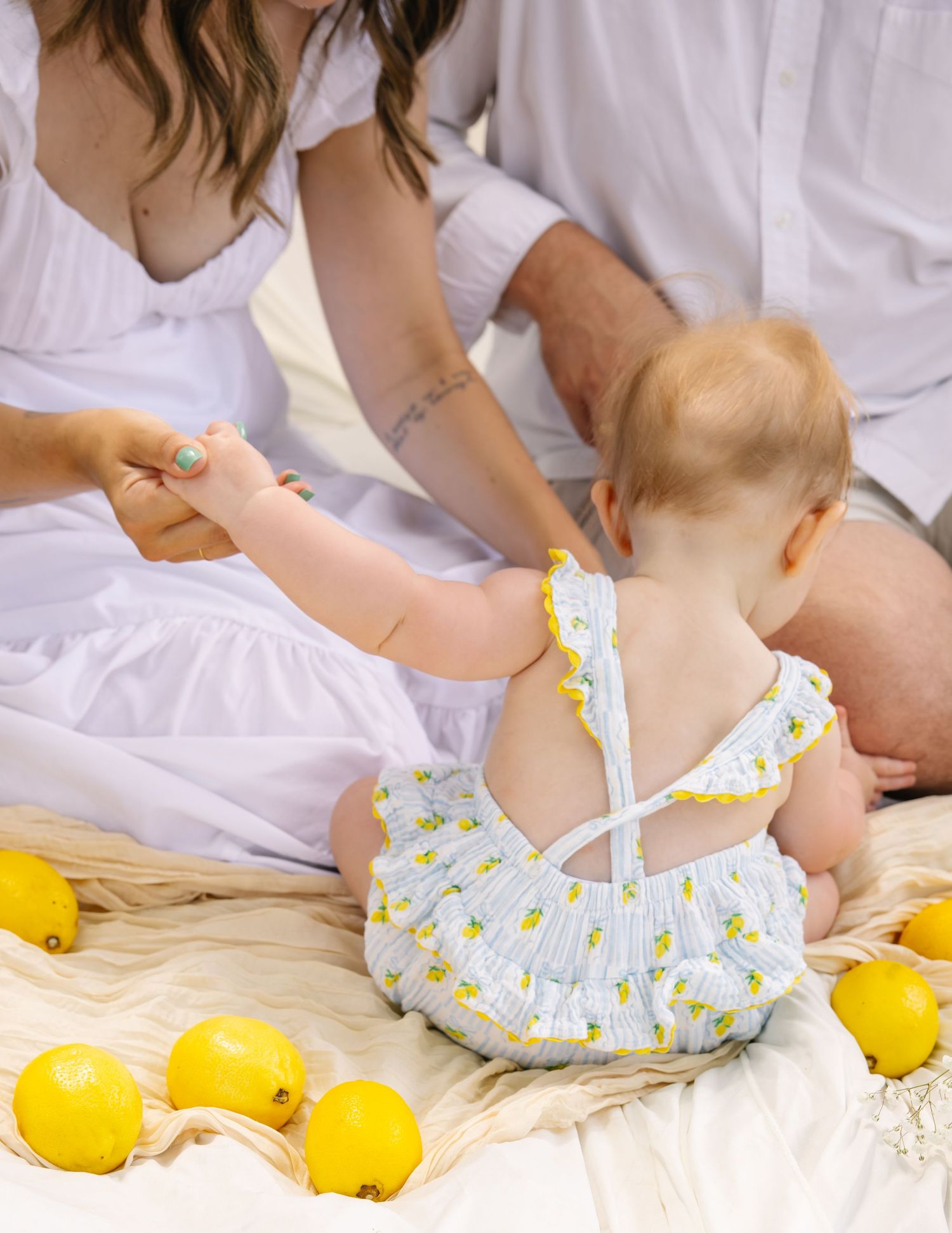 Baby in lemon-print romper reaching toward mother while surrounded by fresh yellow lemons.
