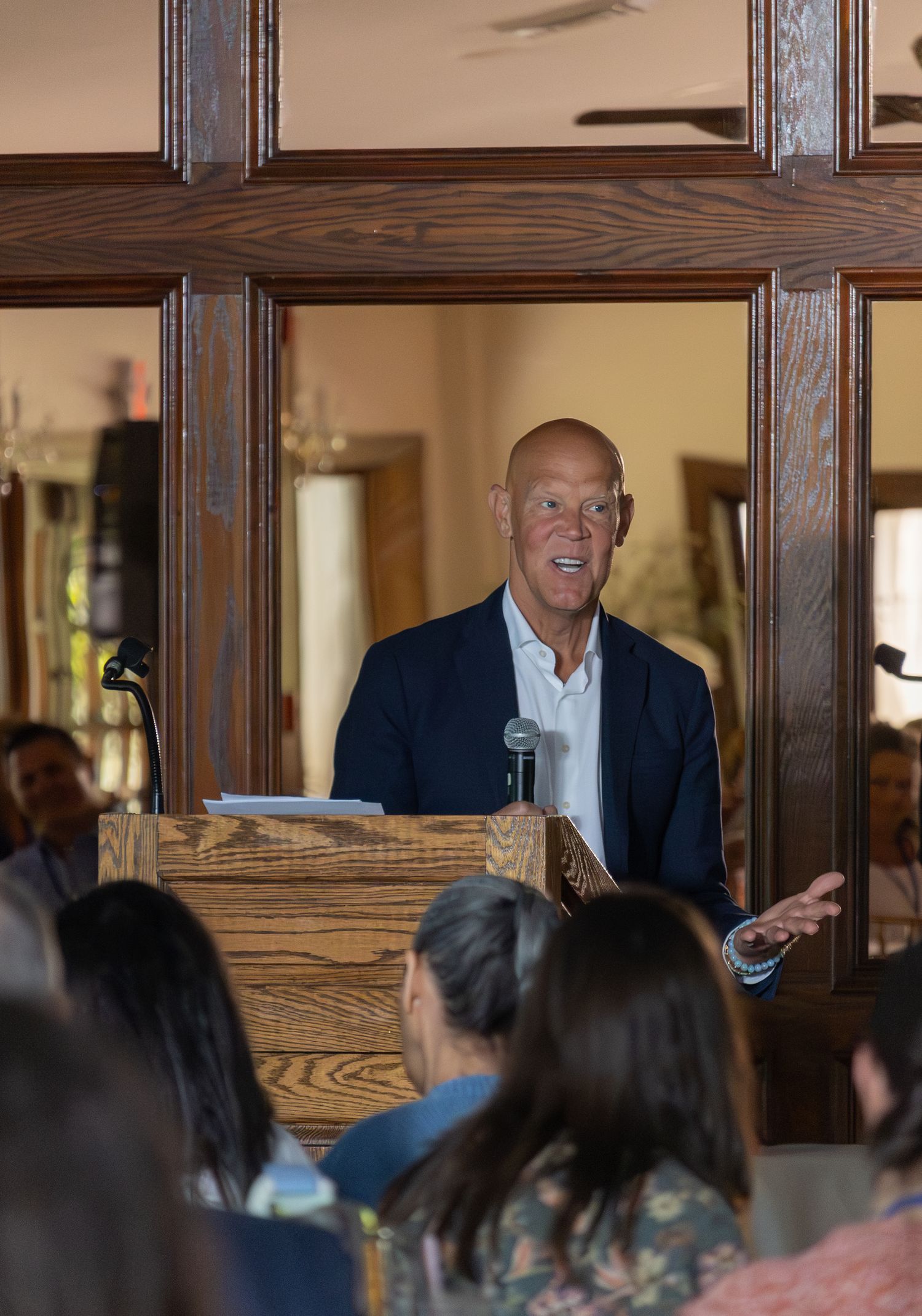 Man in navy blazer speaking at a wooden podium during an indoor conference presentation.