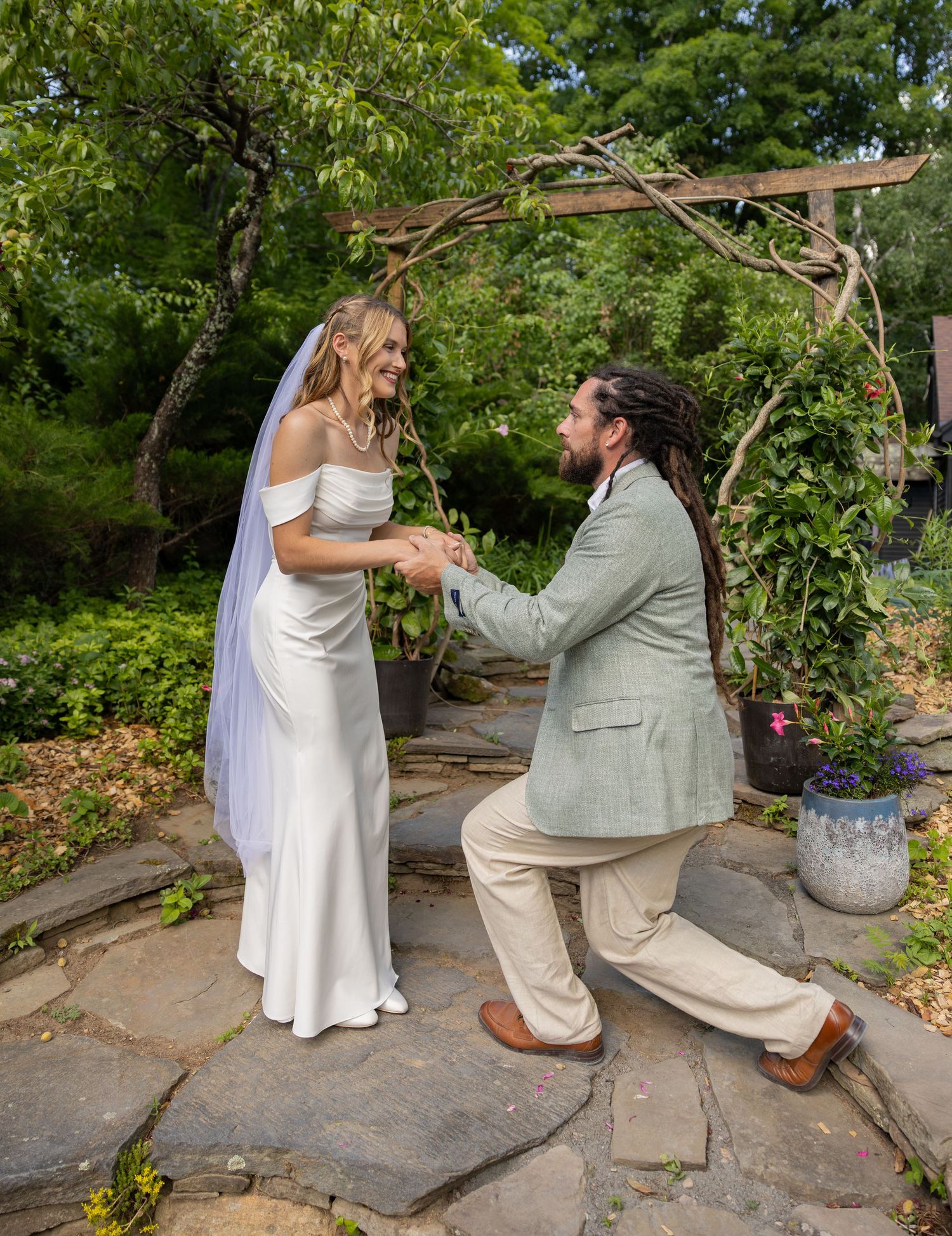 Groom with dreadlocks kneels before smiling bride in white gown under rustic garden arch.