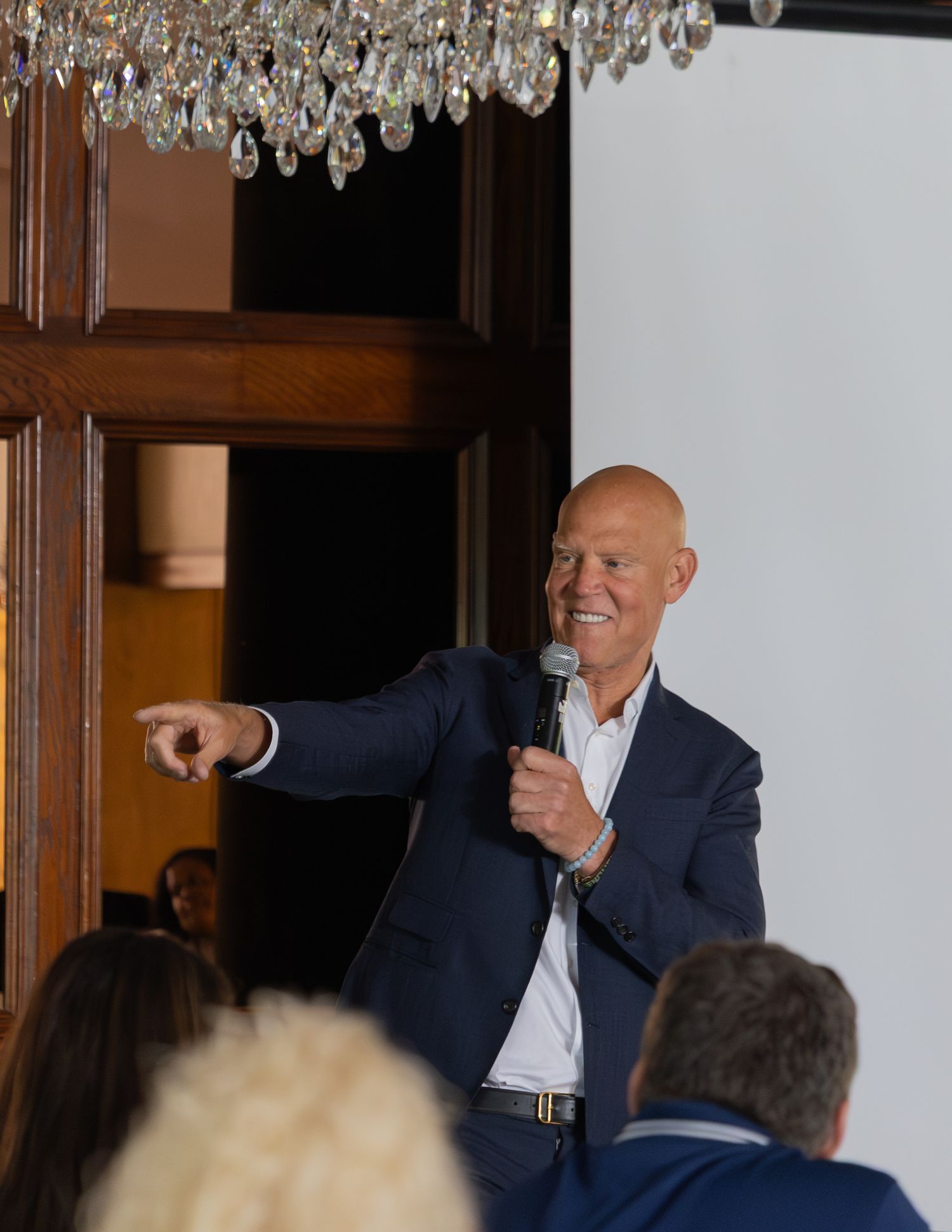 Confident male speaker in navy blazer pointing at audience while holding microphone at indoor event under chandelier.
