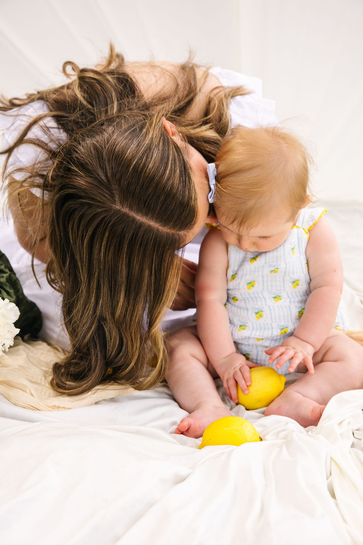 Mother gently kissing her baby holding a yellow lemon on a soft white blanket.