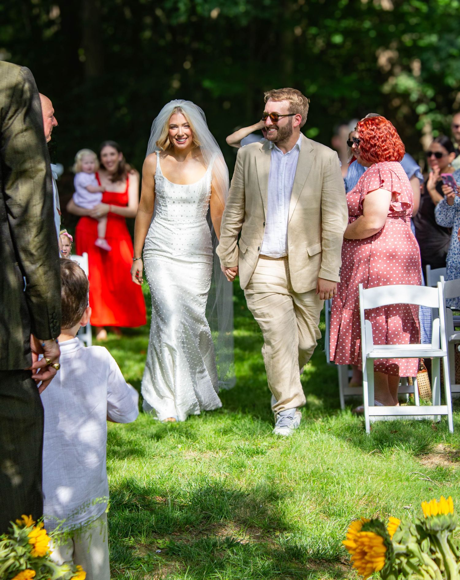 Smiling bride in white gown and groom in tan suit walk down aisle at sunny outdoor garden wedding.