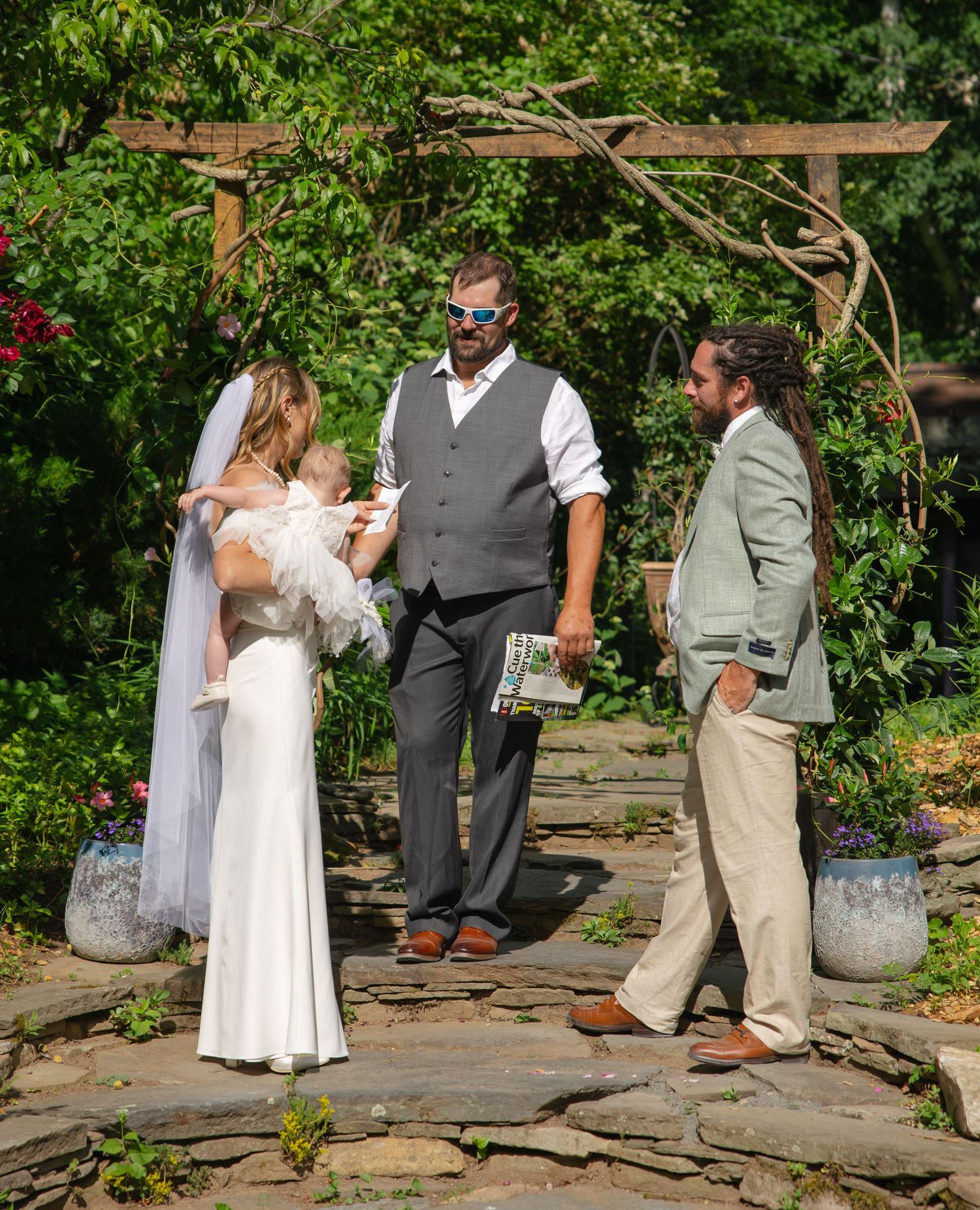 Bride holding infant and groom standing with officiant during intimate outdoor wedding ceremony.