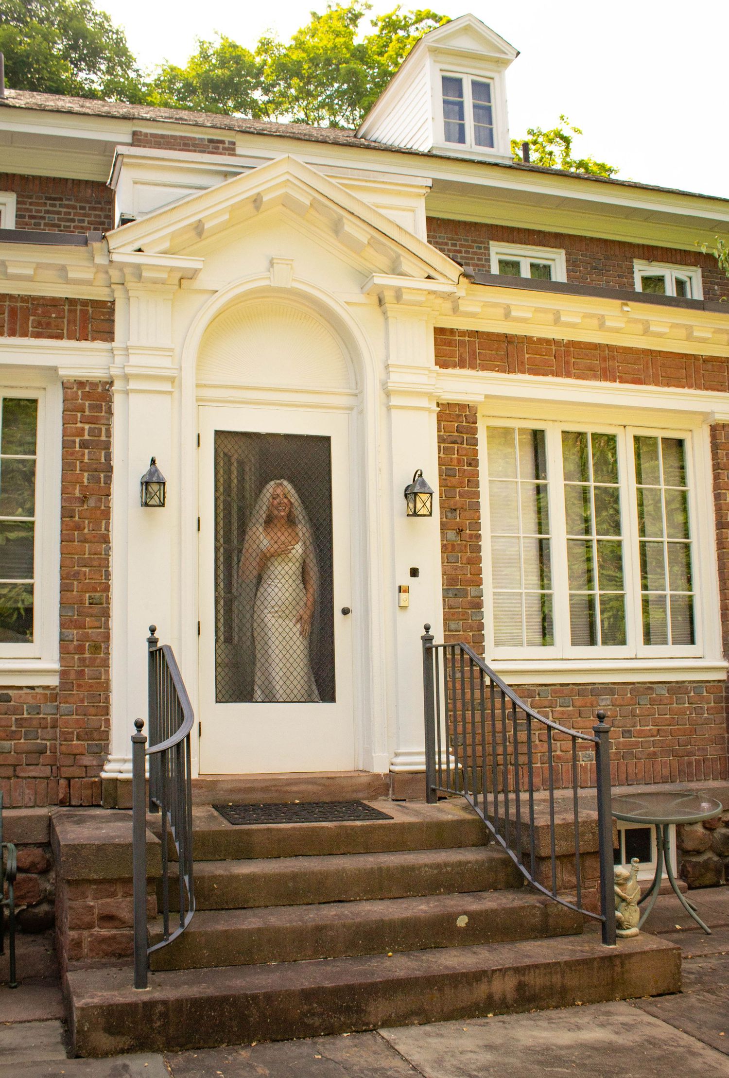 Bride in white gown and veil stands behind glass door of elegant brick home before wedding ceremony.