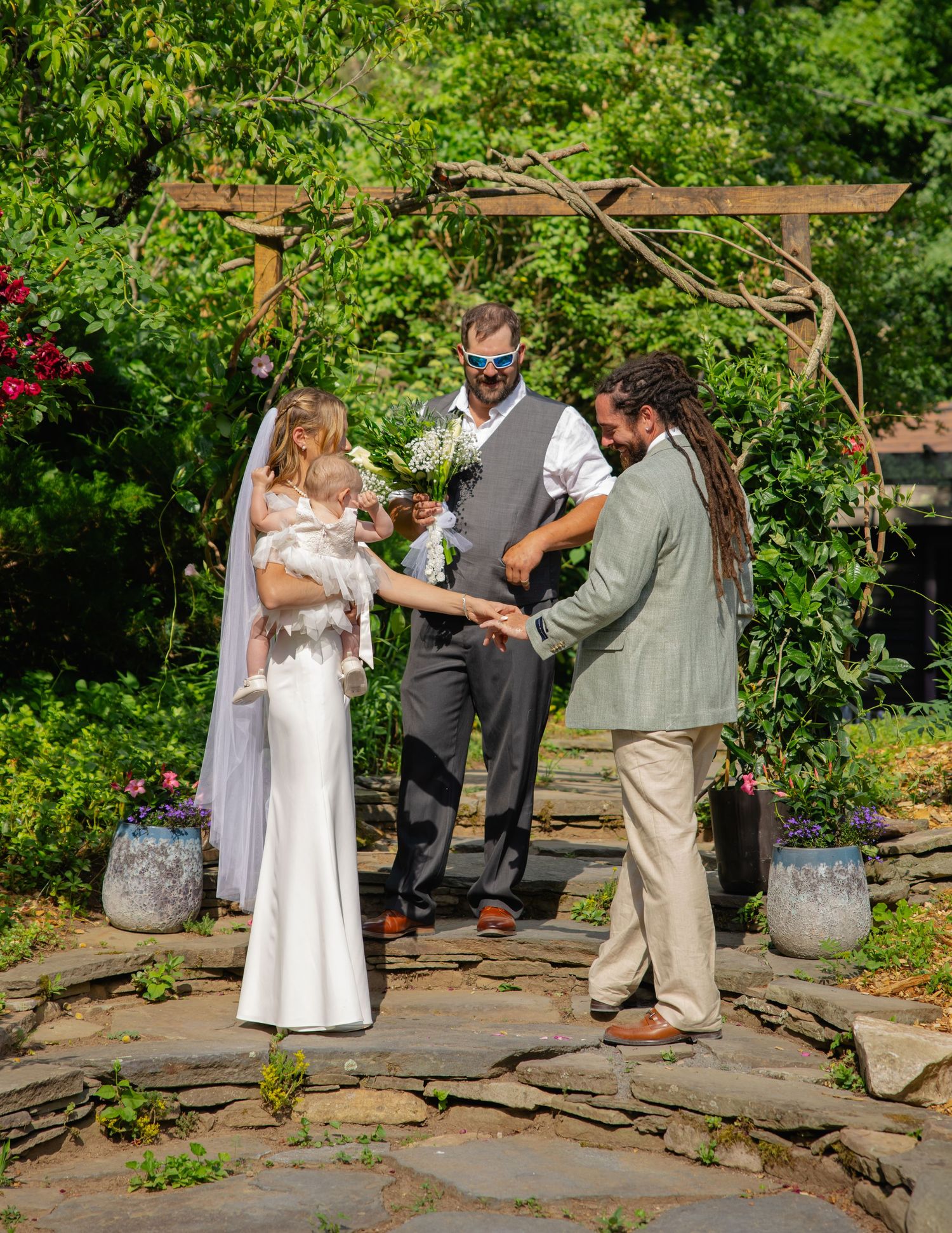 Bride and groom exchanging vows under a wooden arch at a sunny outdoor garden wedding.