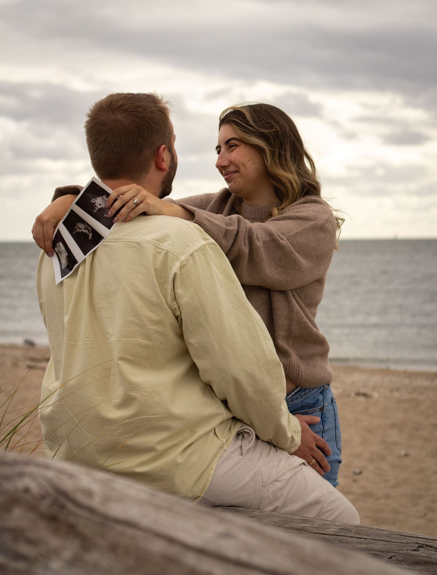 Pregnant woman smiling at partner holding ultrasound photos while sitting together on a beach log.