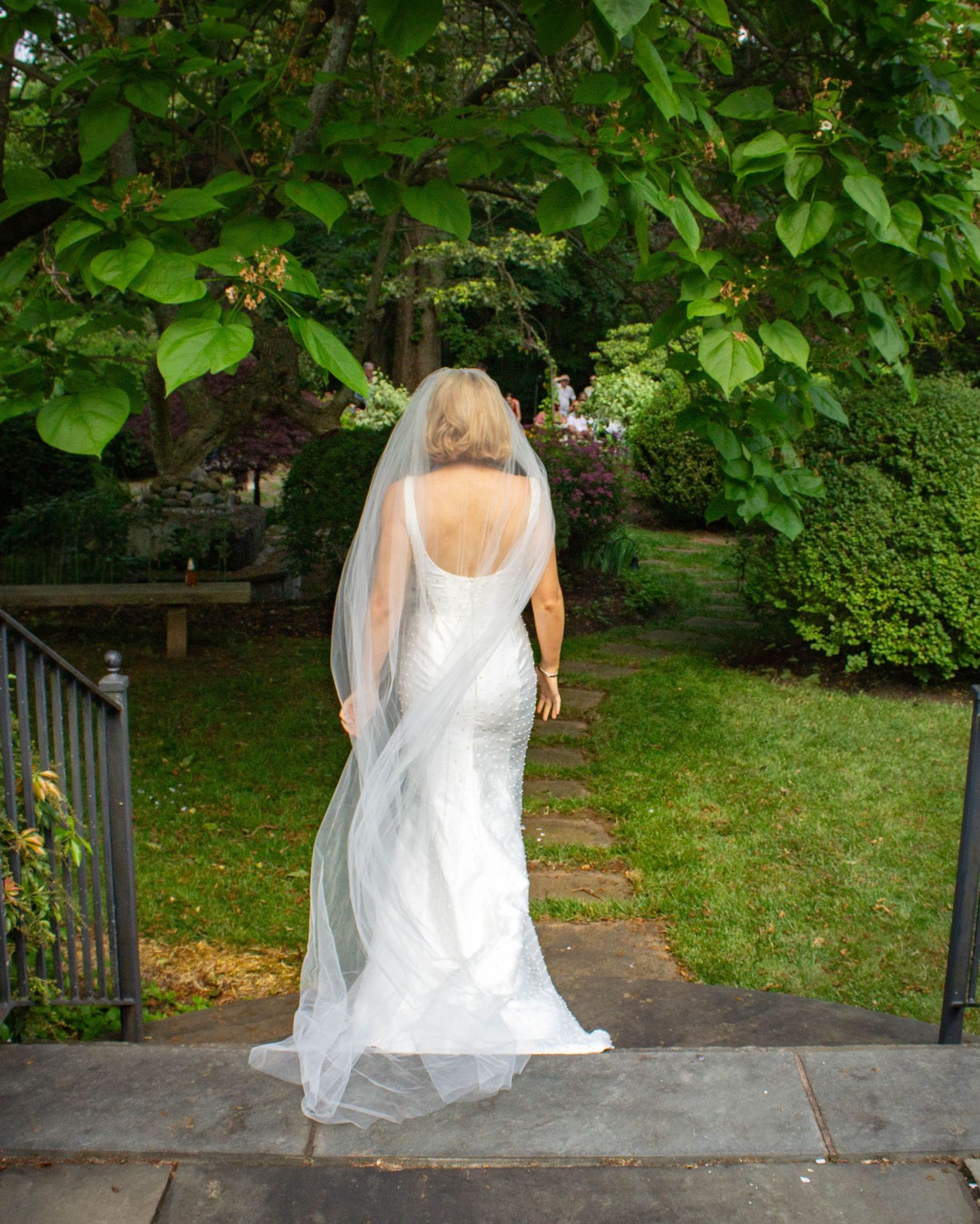 Bride in elegant backless white wedding gown with veil walking through a lush green garden.