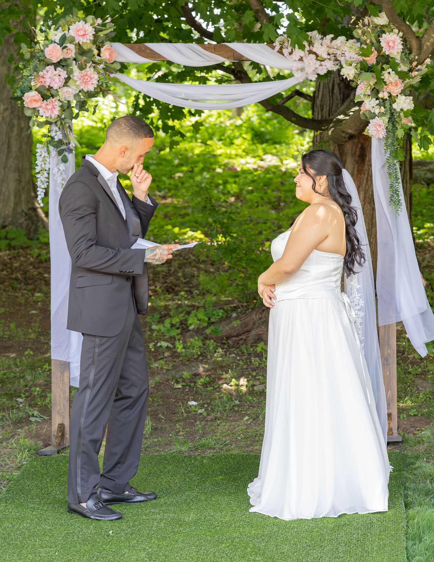 Couple exchanges vows under floral arch with pink blooms during outdoor garden wedding ceremony.
