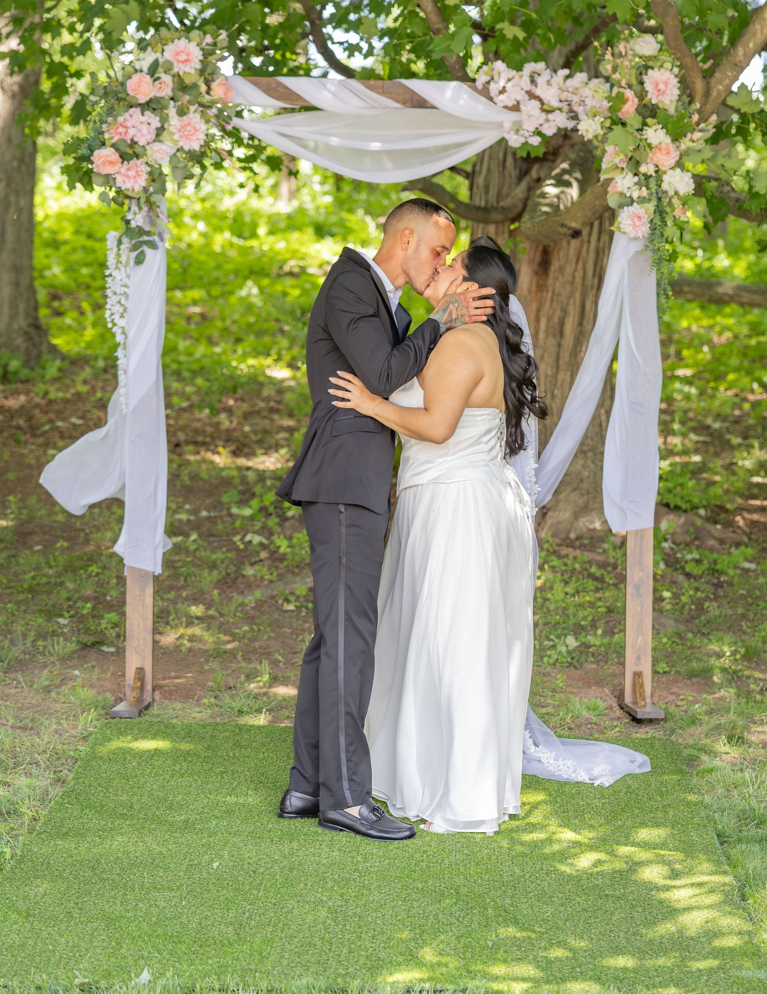 Bride and groom sharing first kiss under floral arch decorated with pink roses at outdoor wedding.