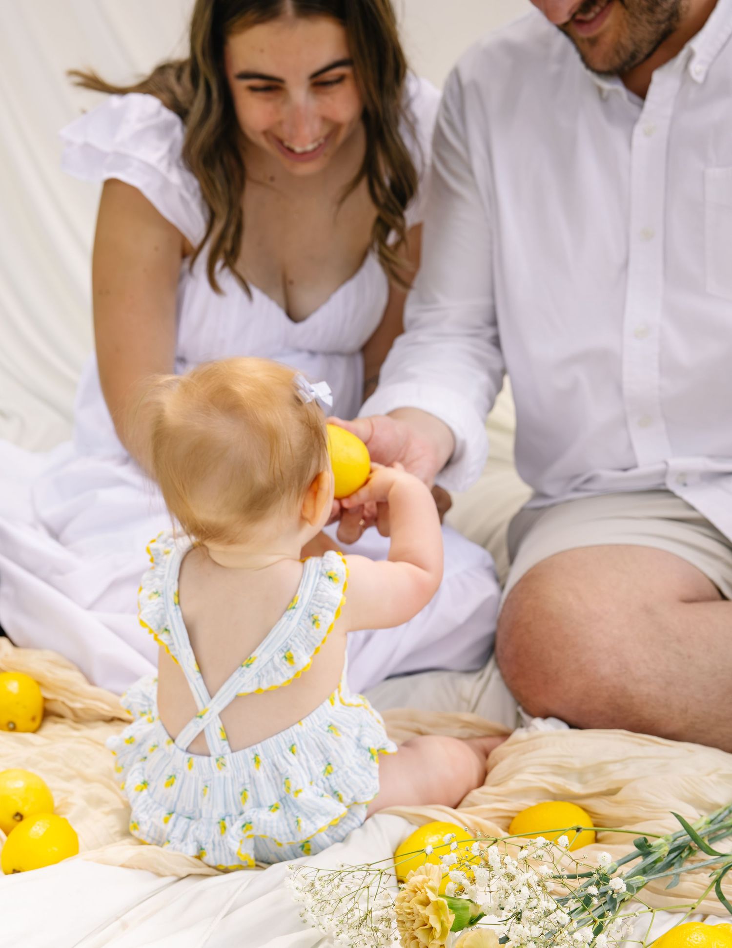 Baby in lemon-print romper playing with a lemon while parents smile warmly beside her.