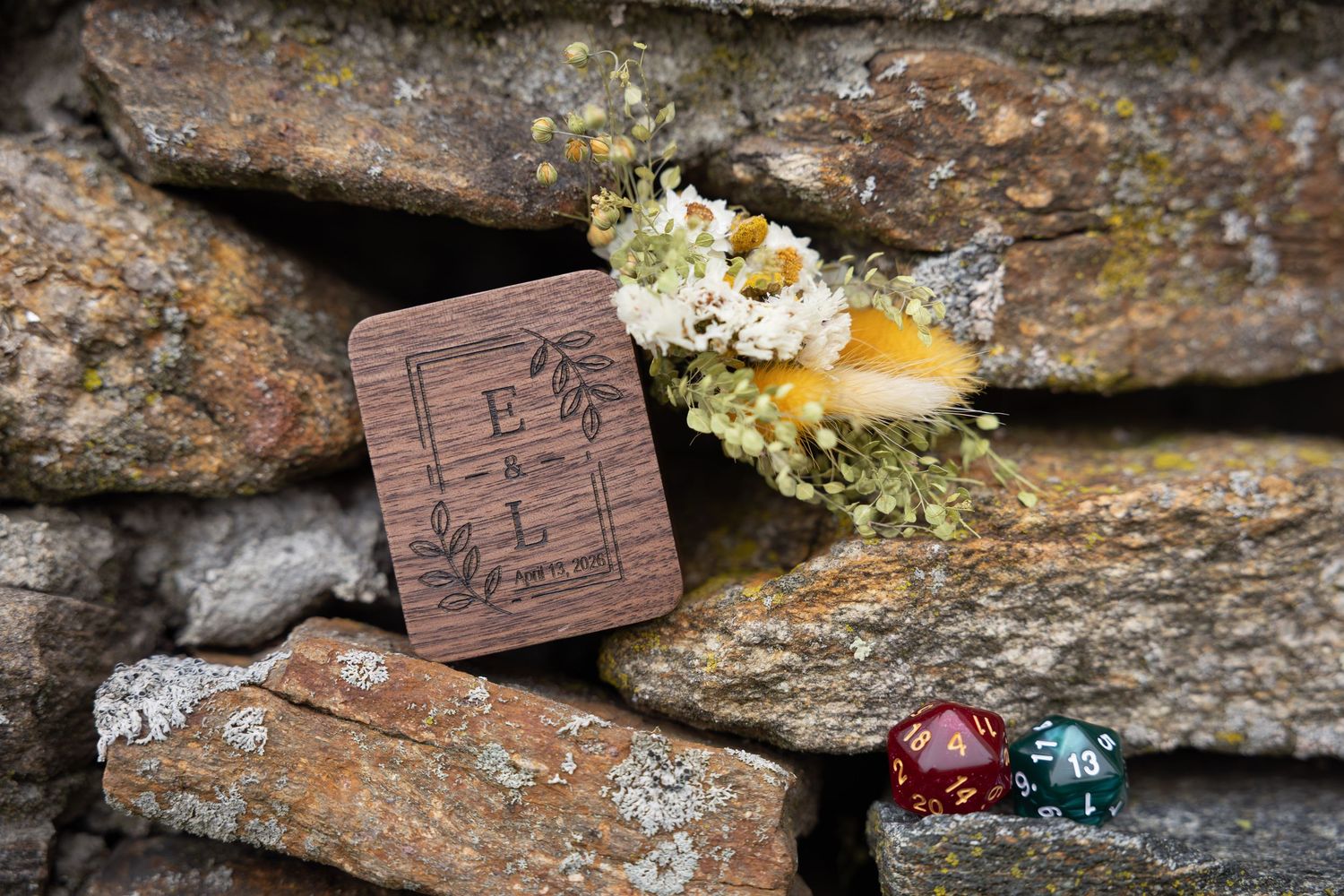 Rustic wooden engraved wedding ring box with floral boutonniere and dice on stone wall.