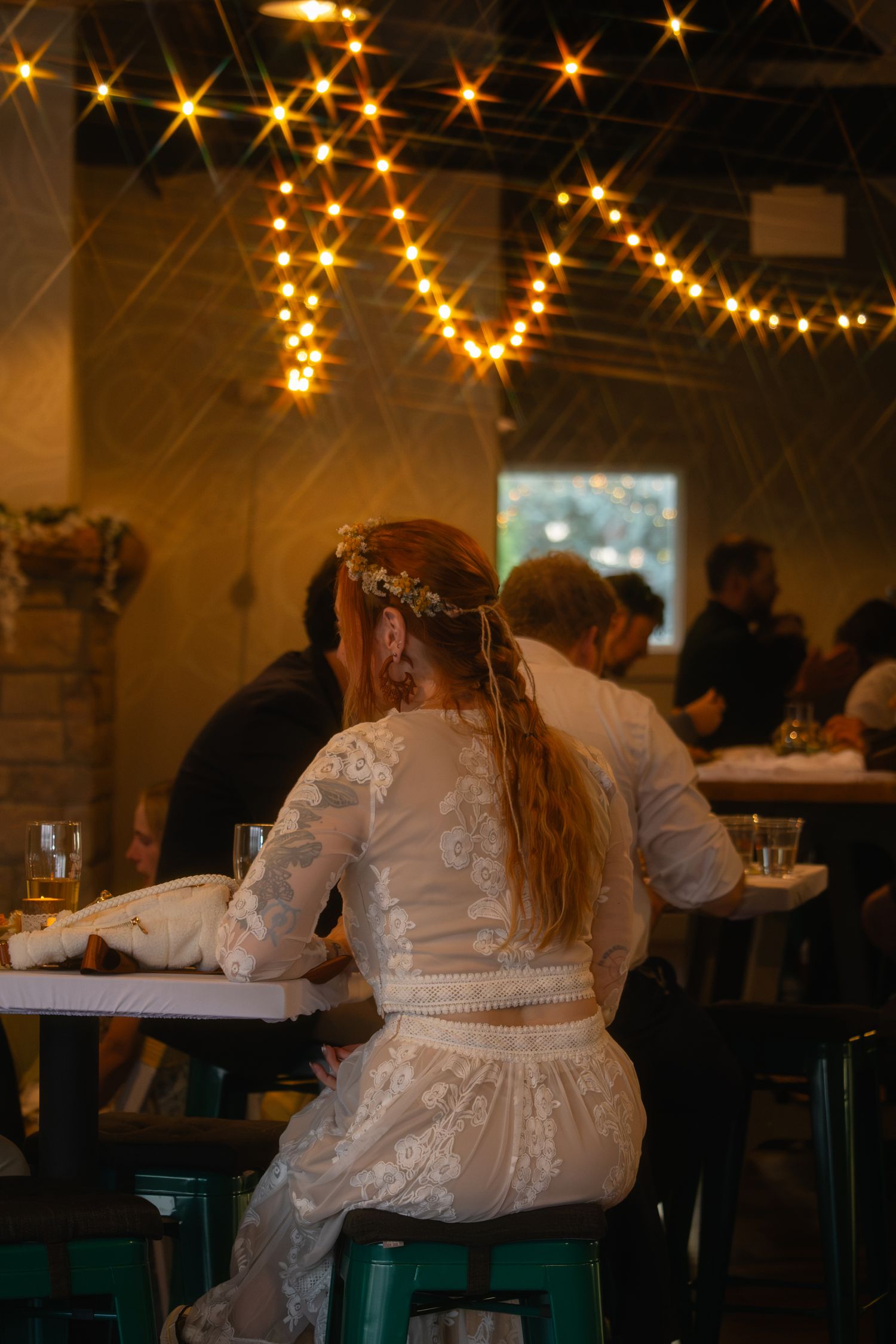 Bride in lace two-piece boho wedding dress with floral crown sits at table under warm string lights.