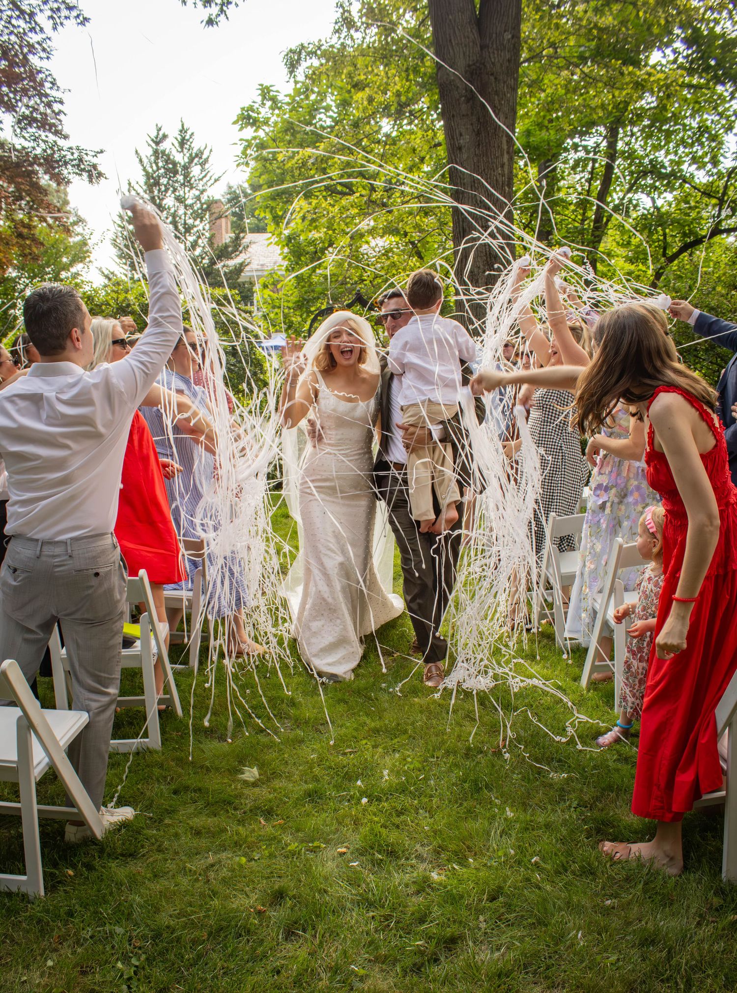 Smiling bride and groom walk through a tunnel of white streamers thrown by cheering guests at outdoor wedding reception.