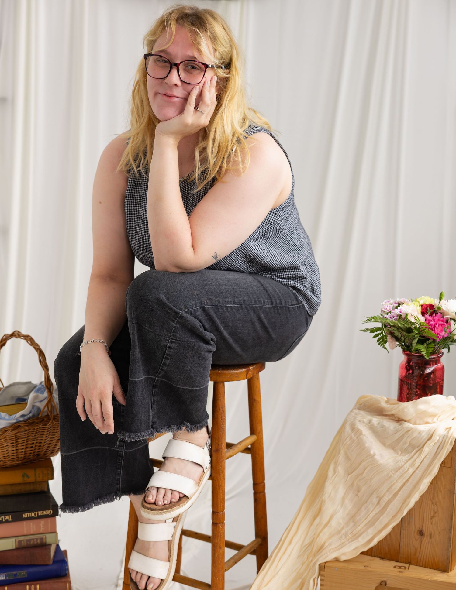 Woman with glasses sitting on a stool in a studio setting wearing a gray tank top and white sandals.