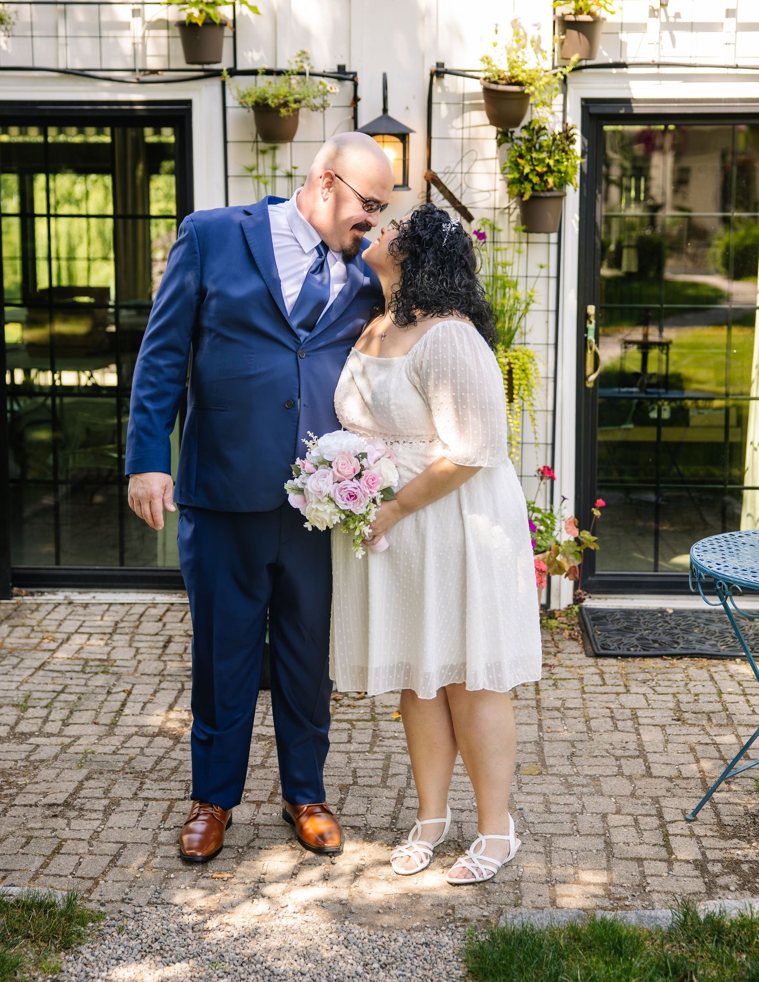 Bride and groom share a romantic moment outdoors, she holds pink bouquet in white dress, he wears navy suit.