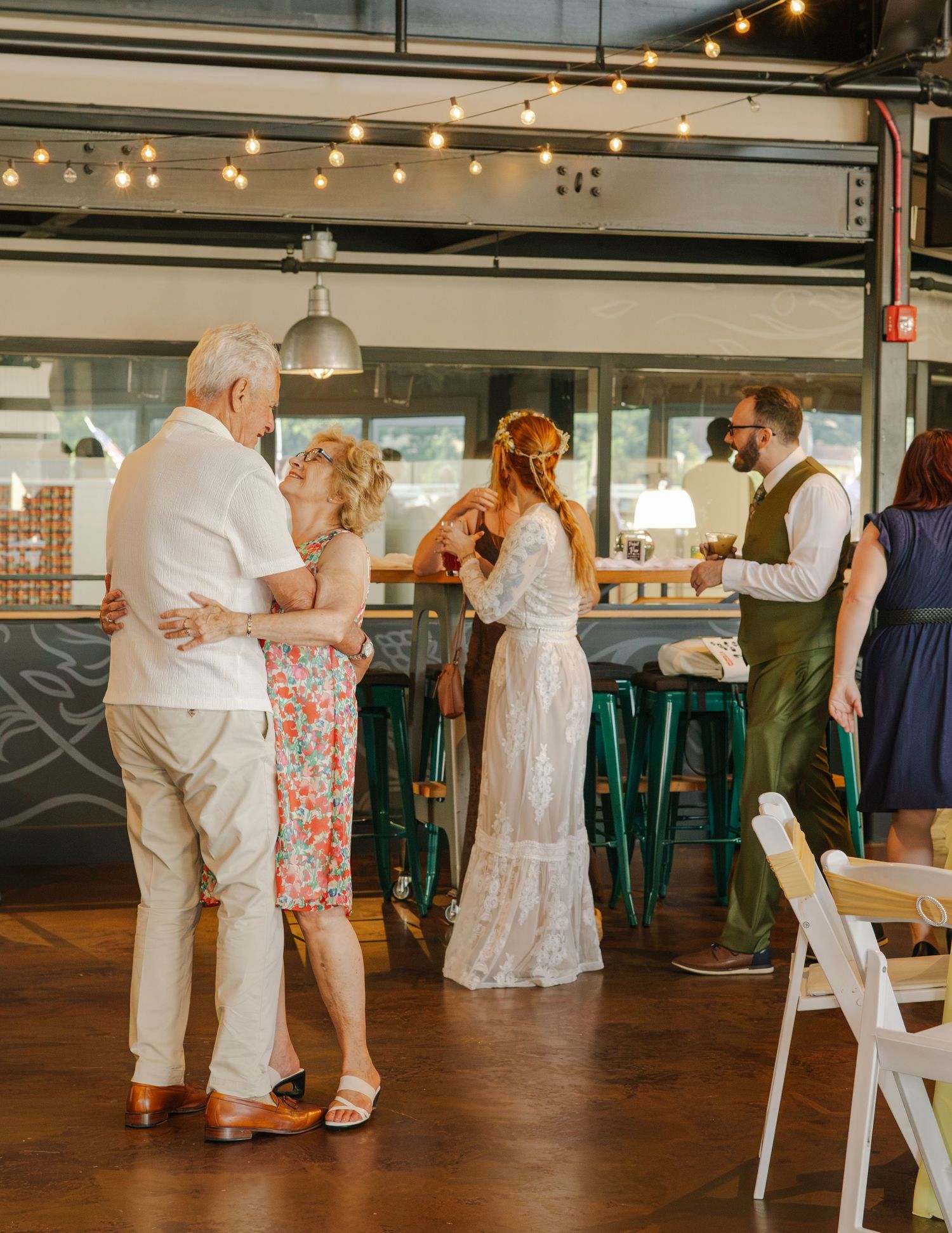 Wedding guests dancing and socializing inside rustic venue with string lights and teal bar stools.