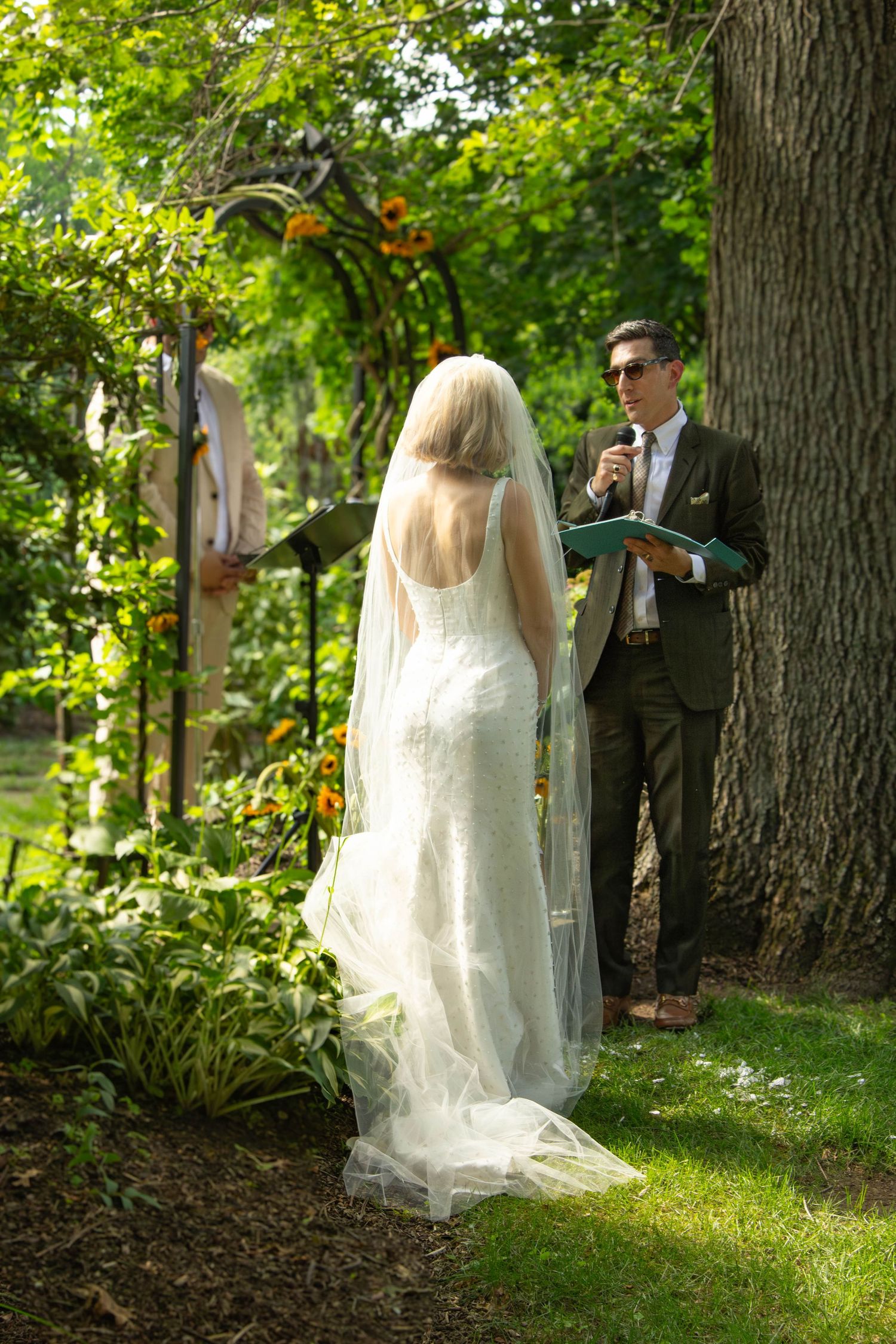 Bride in white gown and veil stands before officiant during intimate outdoor garden wedding ceremony.