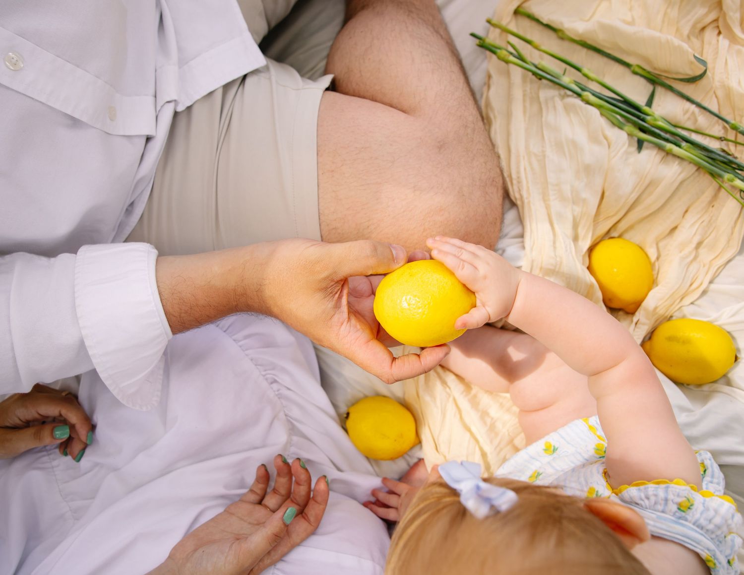 Adult hands passing a yellow lemon to a baby in a lemon-print outfit on a soft blanket.