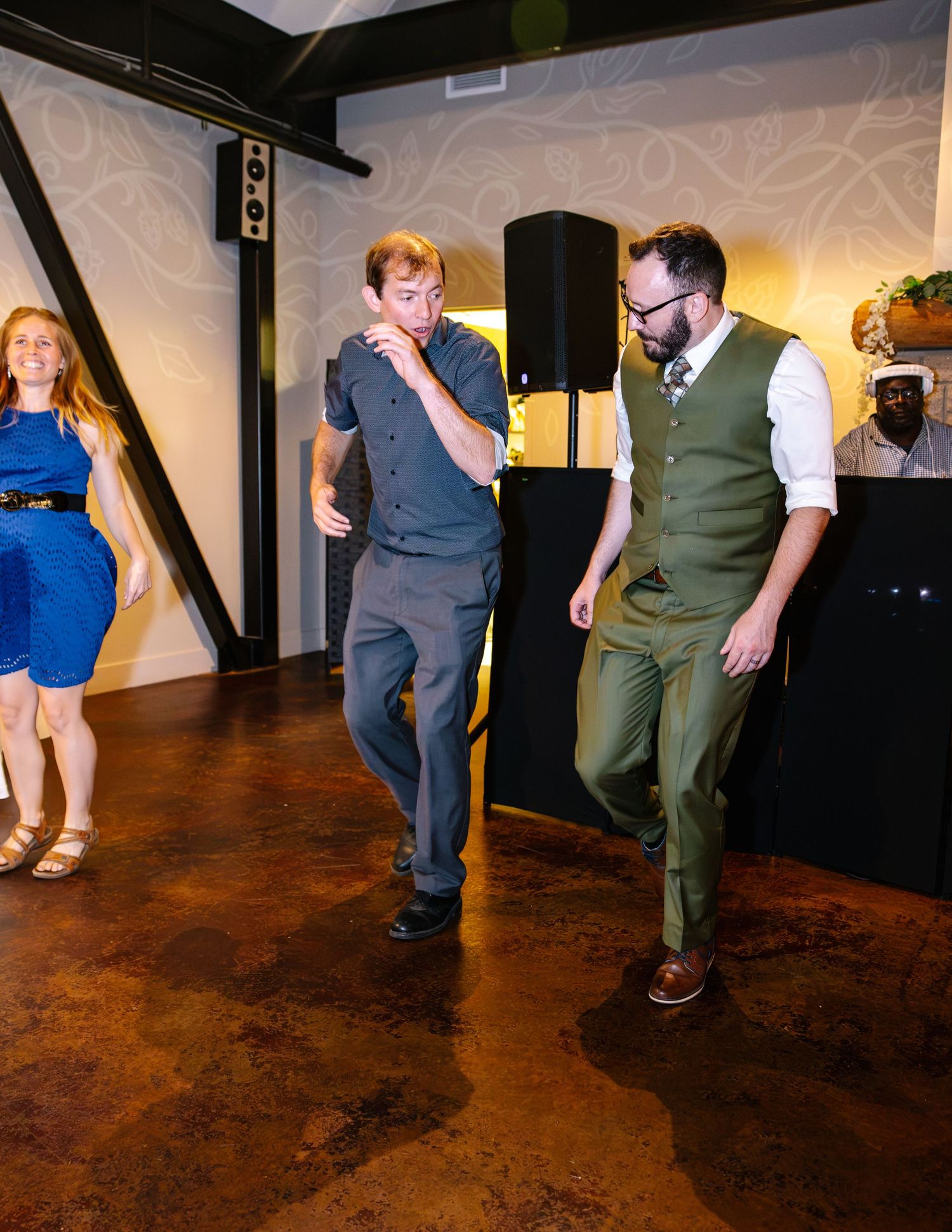 Groom in olive vest and guests dancing energetically on polished wood dance floor at wedding reception.