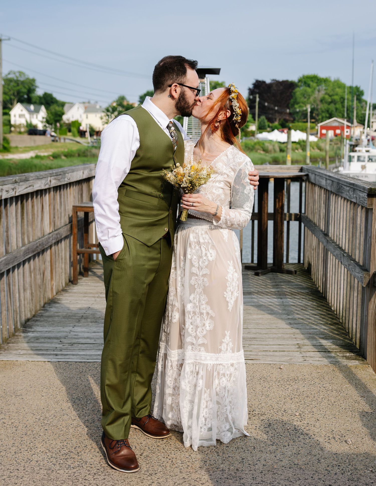 Bride and groom share a kiss on a wooden dock, she in lace gown with floral crown, he in olive suit.