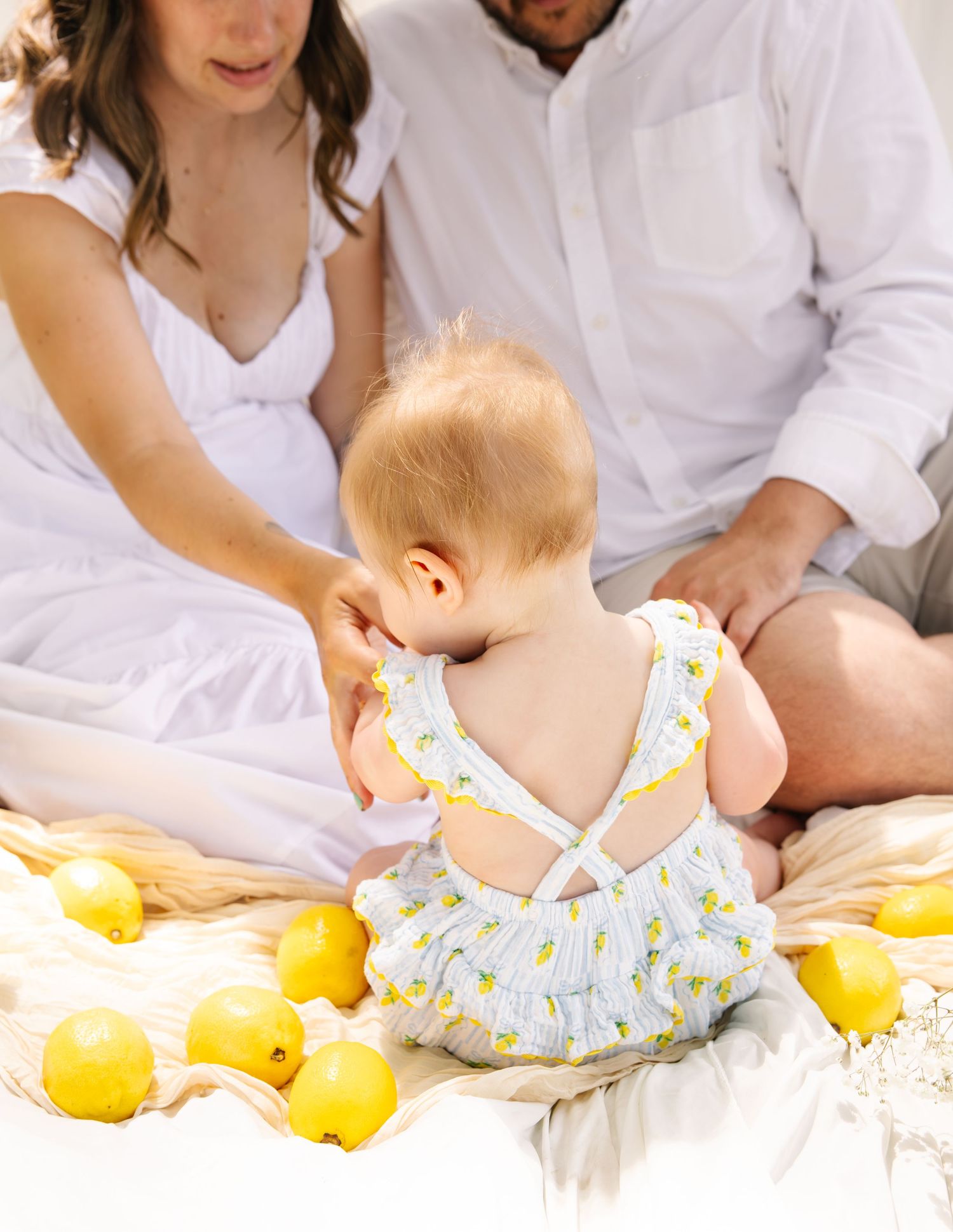 Back view of baby in lemon-print romper sitting between parents on a white blanket.