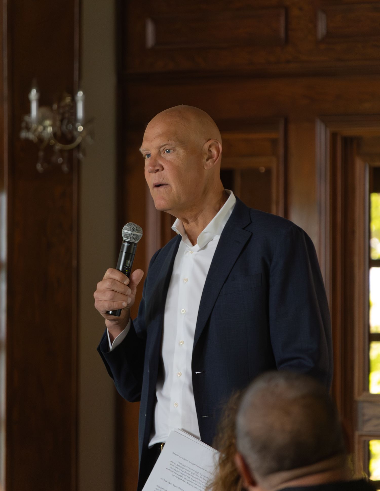 Bald man in navy blazer holds microphone and papers while speaking at wood-paneled indoor event.