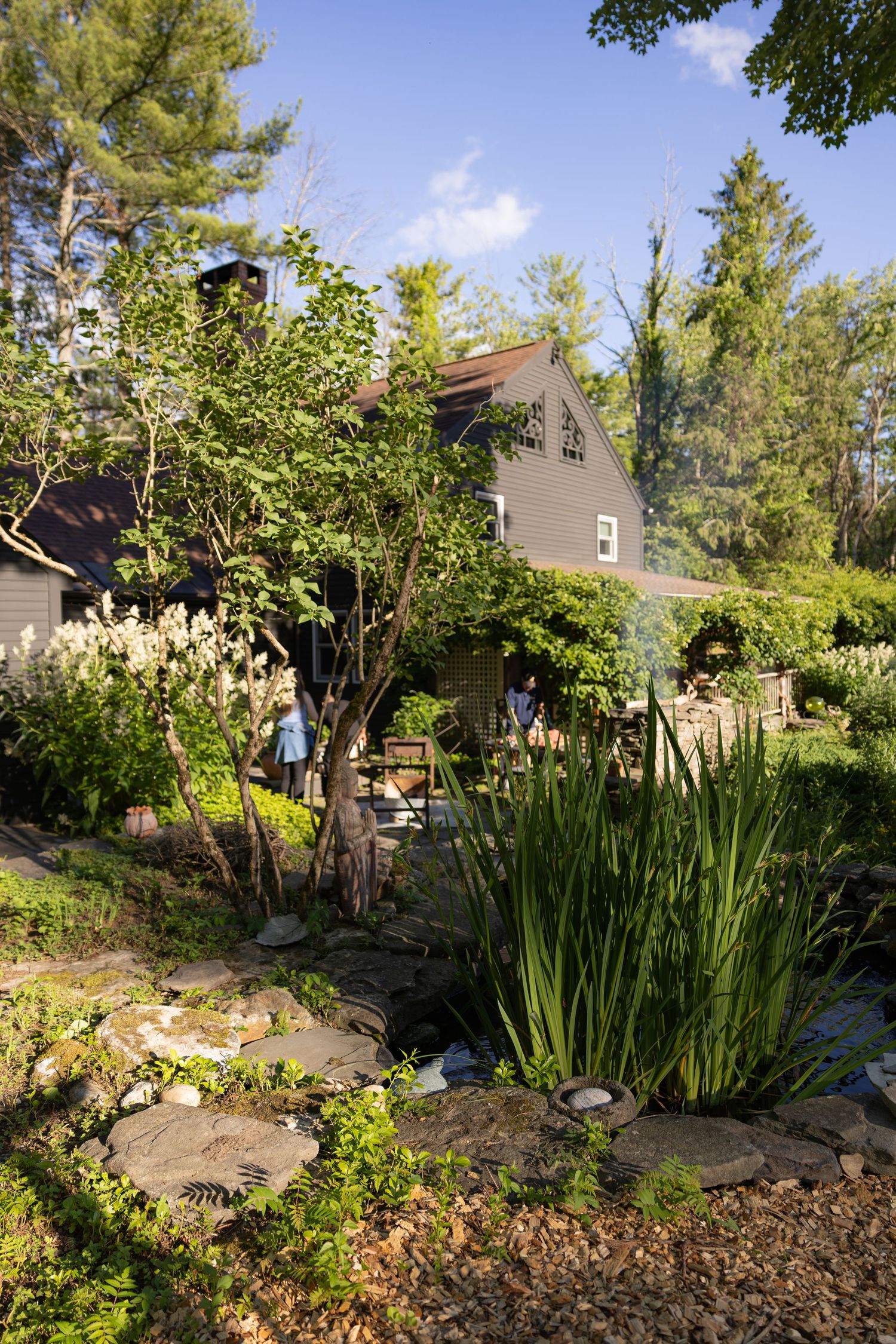 Scenic New England farmhouse surrounded by vibrant green gardens with stone pathway and pond.