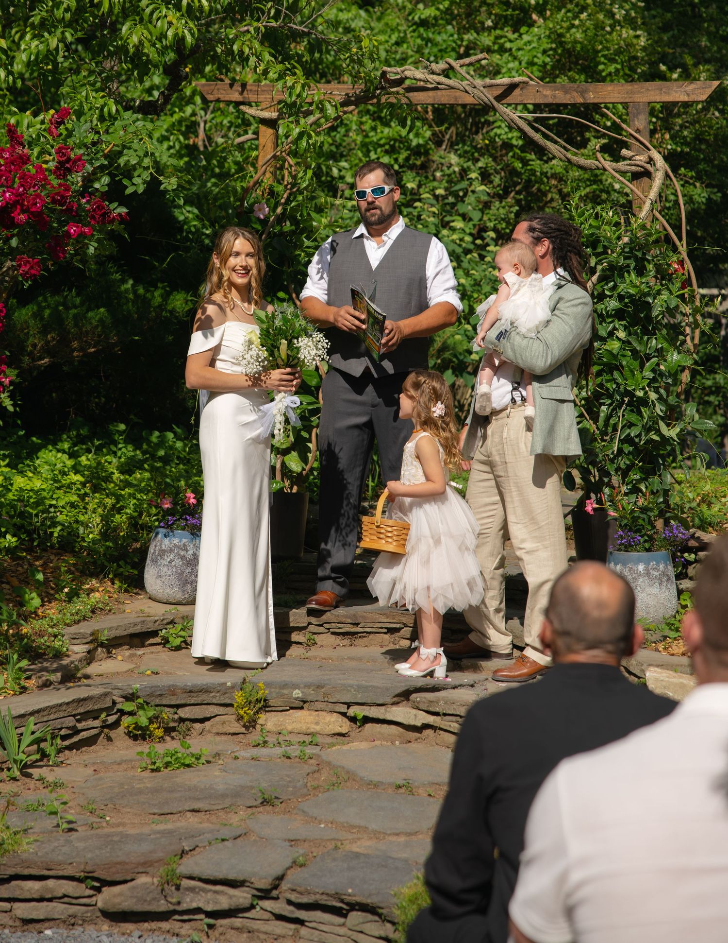 Bride, groom, and flower girl standing with officiant during a sunny outdoor garden wedding.
