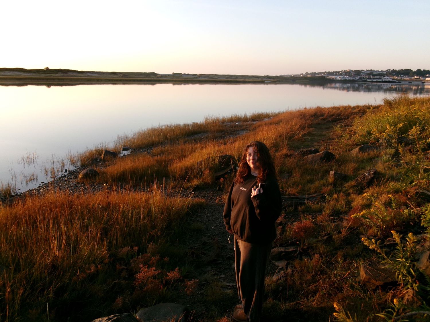 Woman standing on rocky shoreline beside golden marsh grass at sunset with calm water reflection.