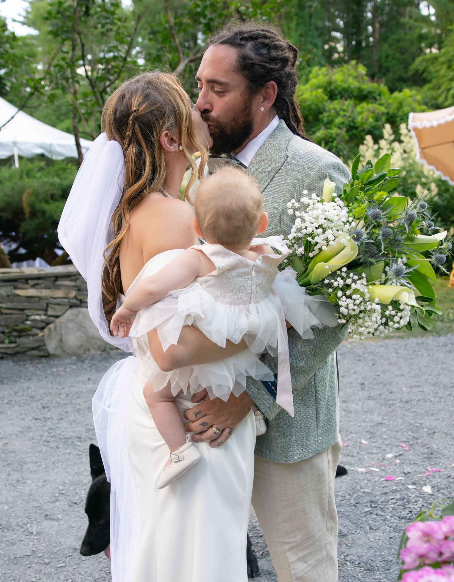 Newlywed couple sharing a kiss while holding their baby at an outdoor garden wedding.