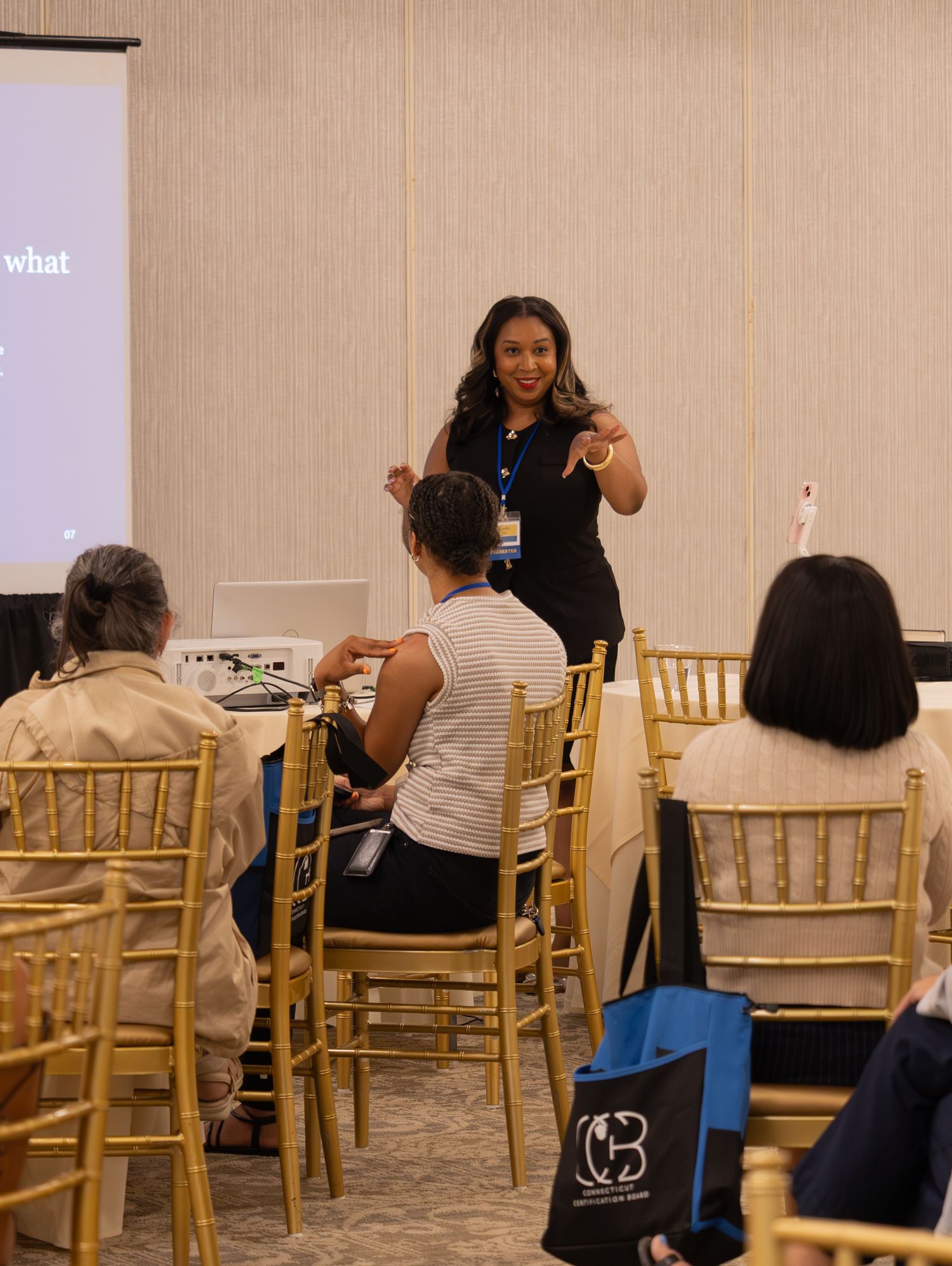 Woman in black outfit presenting to seated audience in a conference room with a projection screen.