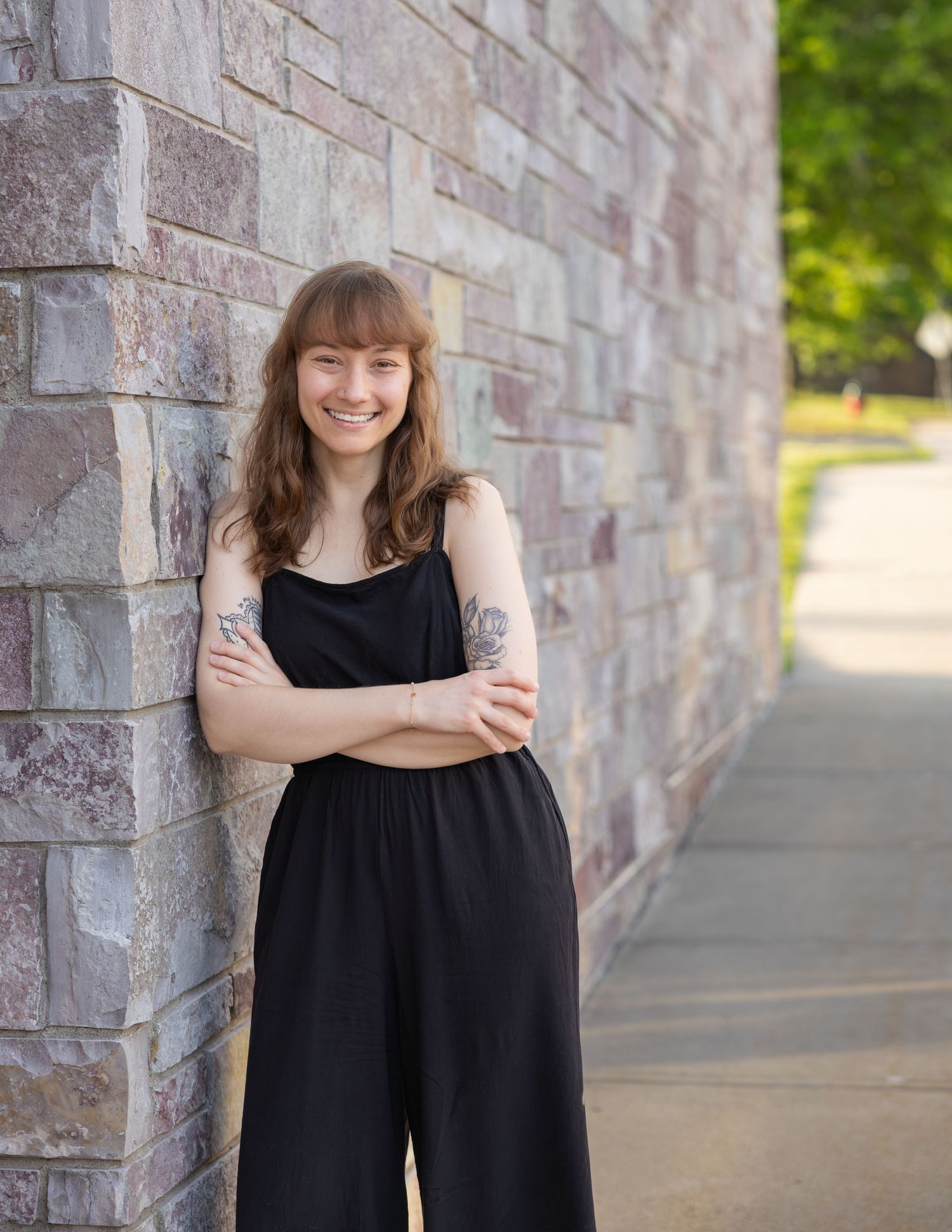 Smiling young woman with tattoos in black jumpsuit leans against brick wall.