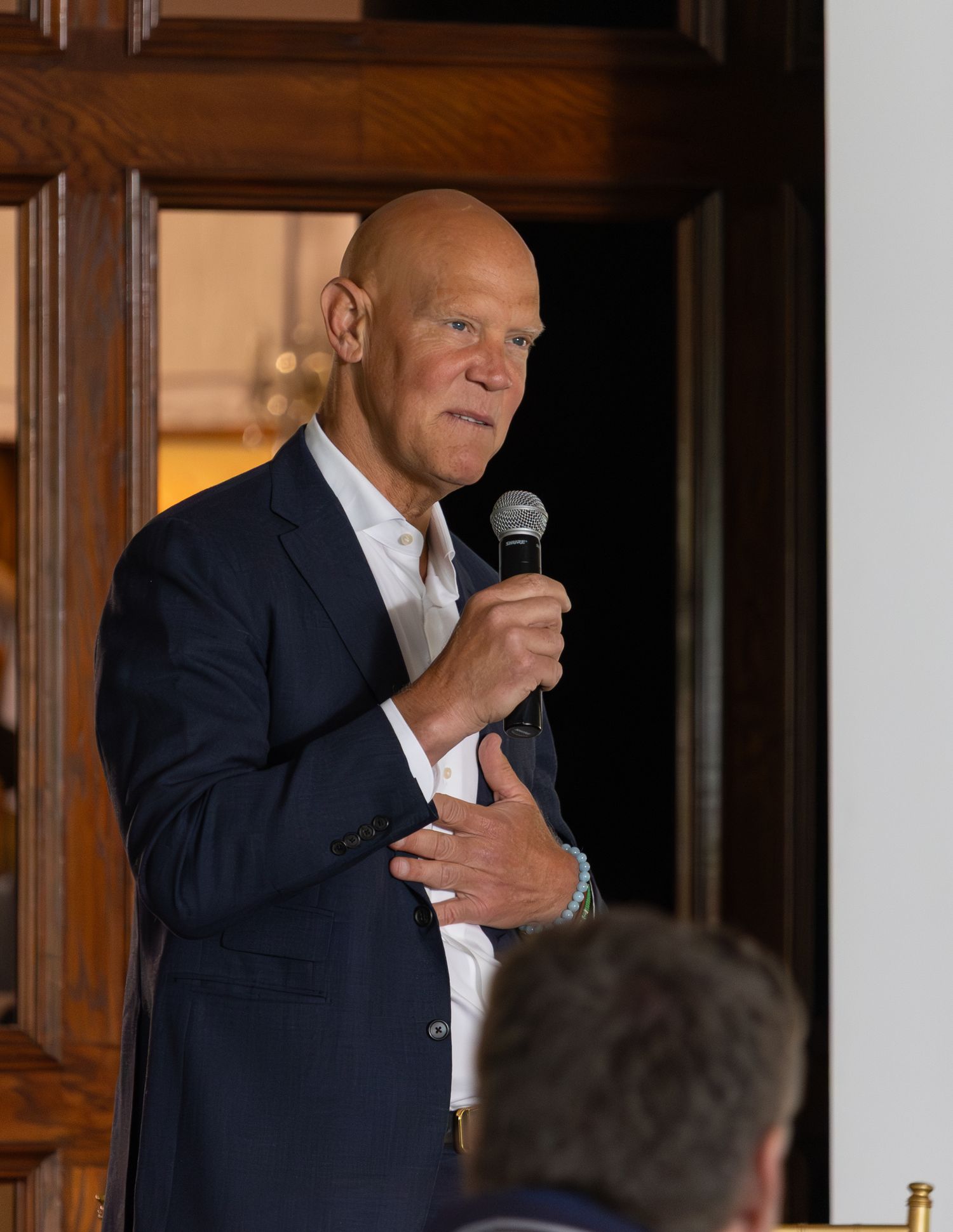 Male speaker in navy blazer holding microphone while addressing an audience at a formal event.