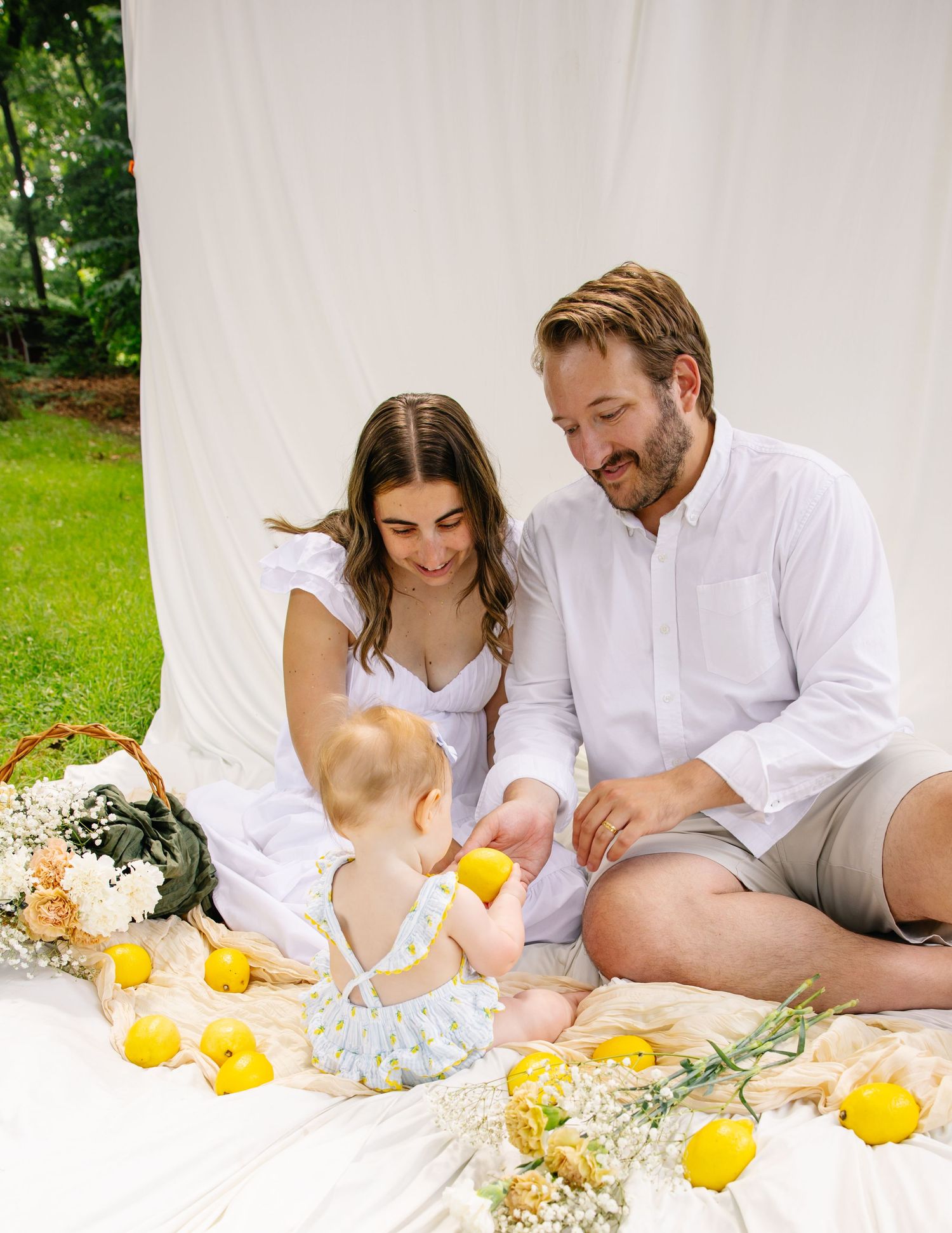 Family of three in white outfits playing with lemons on a white blanket outdoors.