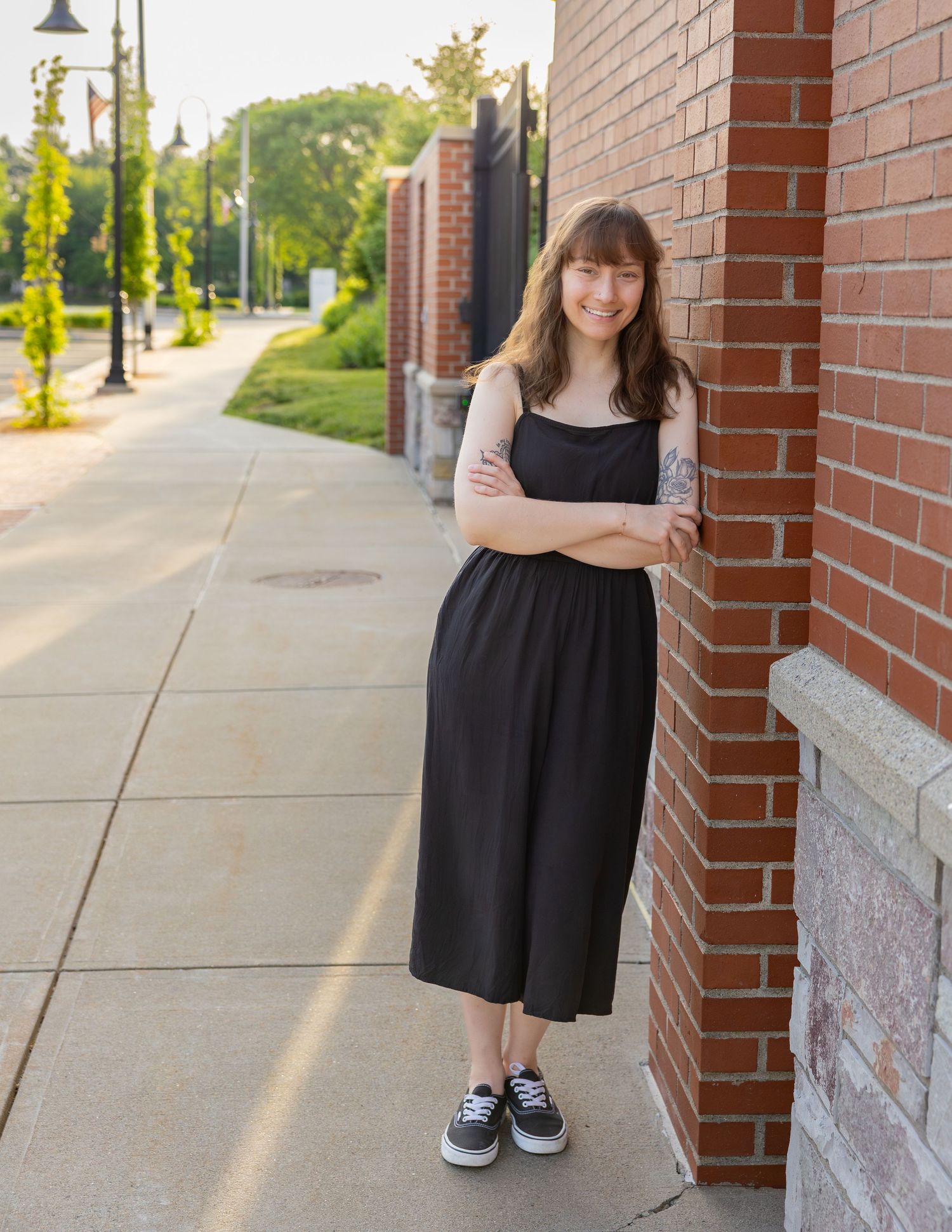 Smiling young woman in black midi dress leans against red brick wall.
