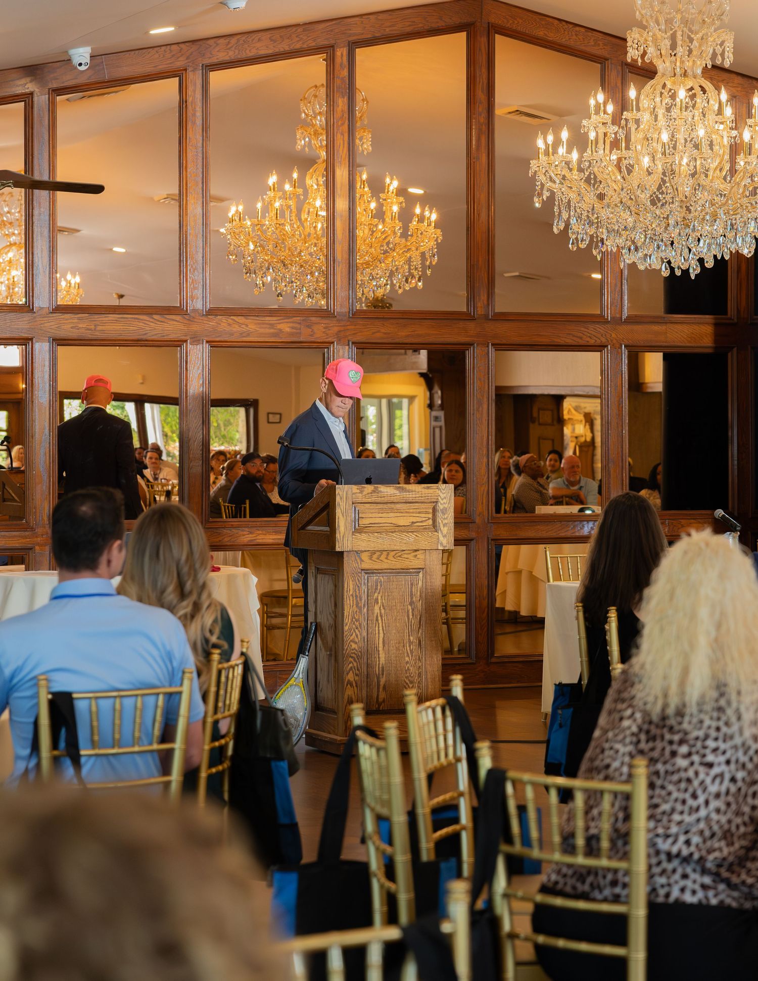 Speaker at podium wearing pink hat addressing seated audience in elegant chandelier ballroom venue.