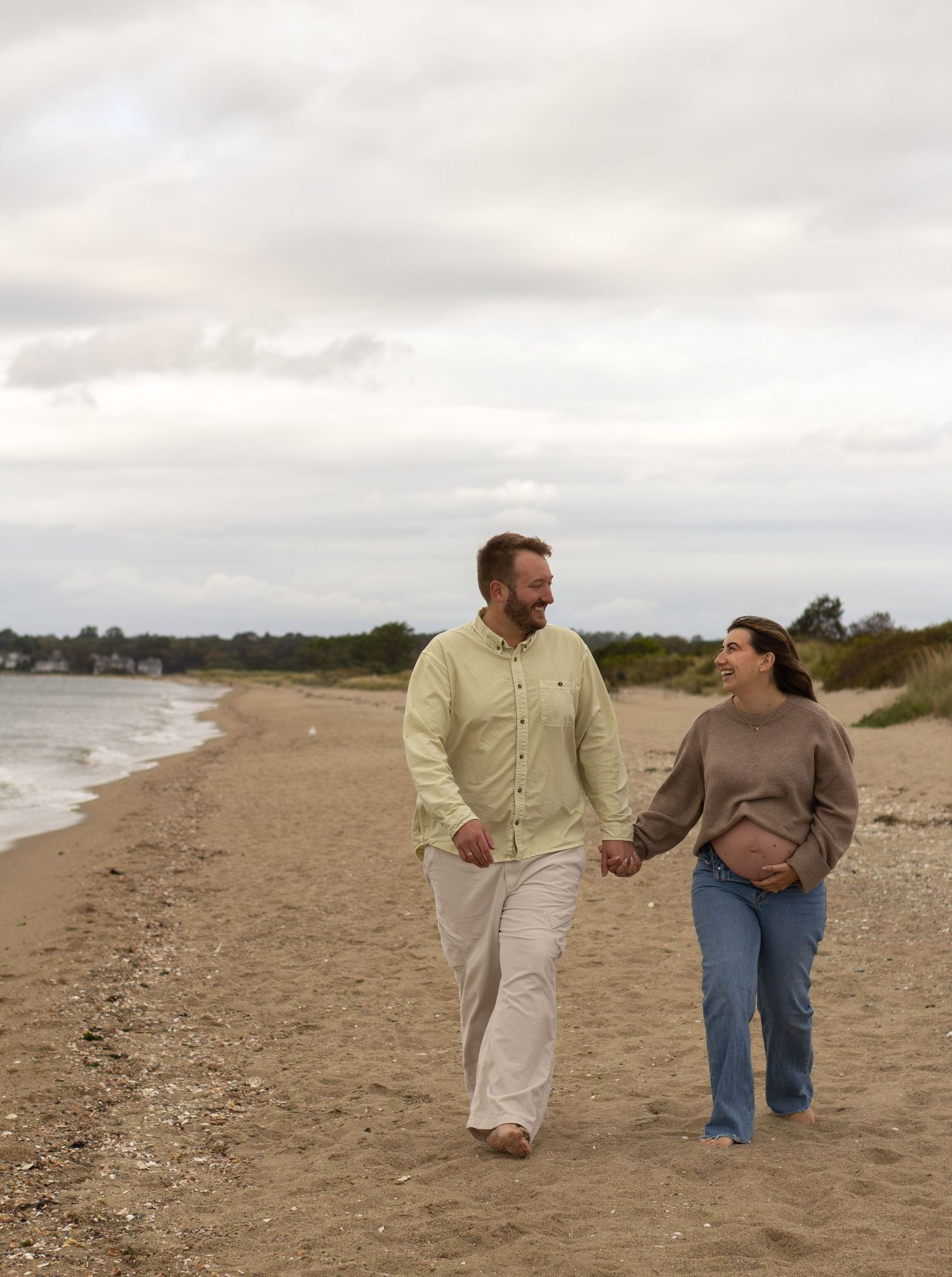 Smiling pregnant couple holding hands and walking along a sandy beach on a cloudy day.