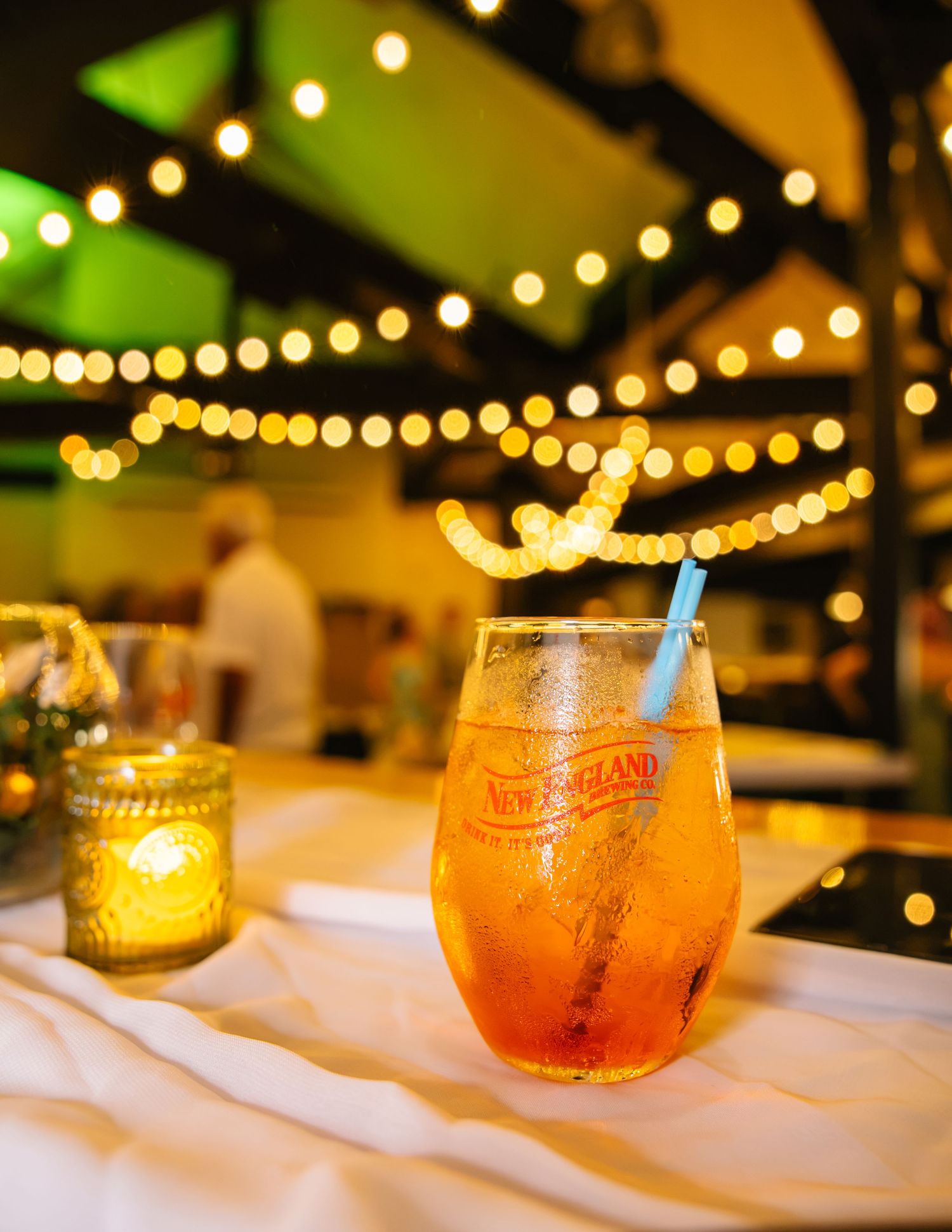 Amber cocktail with a curly straw in a New England branded glass at an event with bokeh string lights.