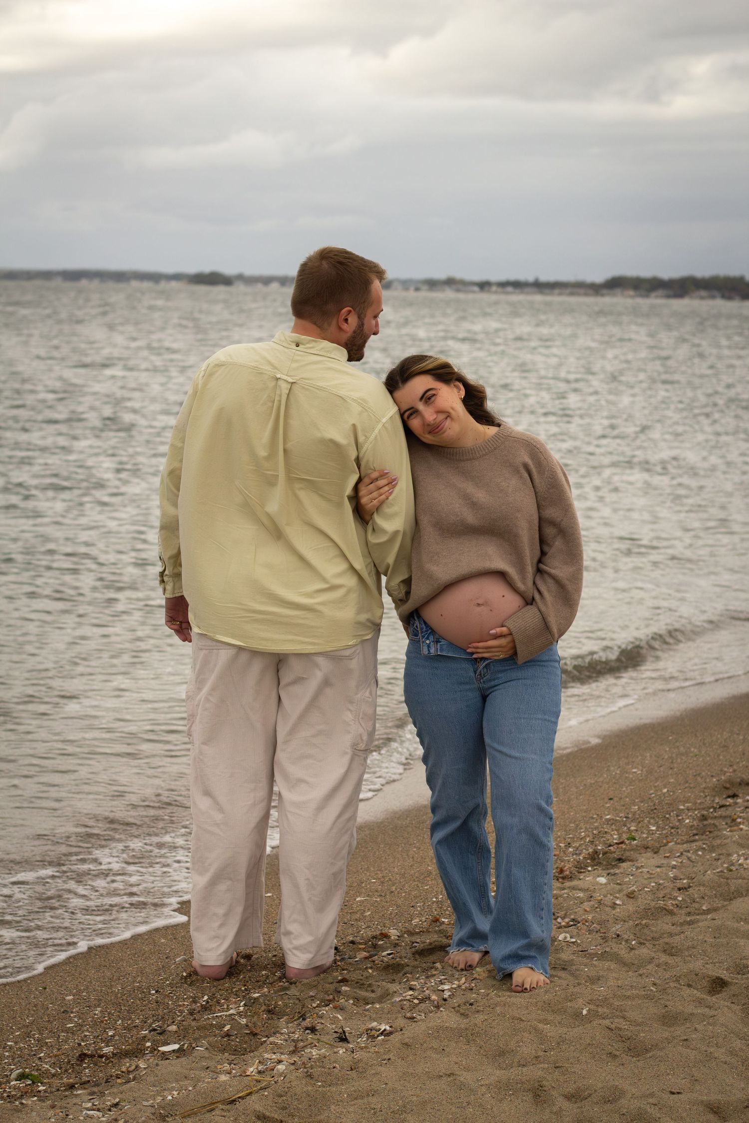 Pregnant woman in brown sweater and jeans leaning on partner while standing on a sandy beach.