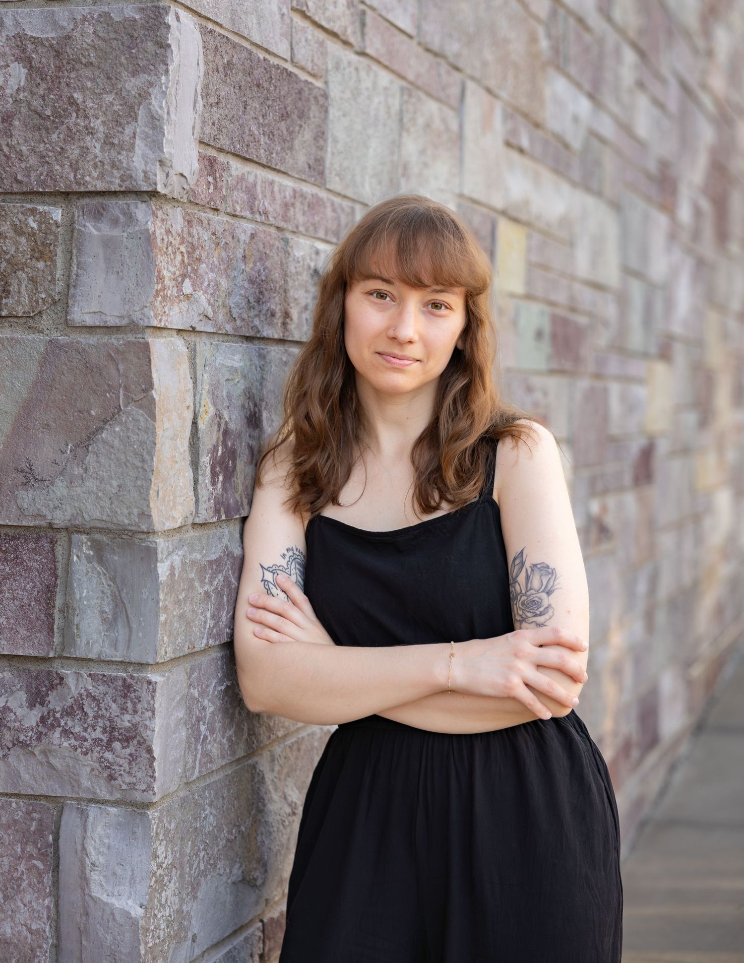 Young woman with tattoos in black dress leaning against stone brick wall outdoors.